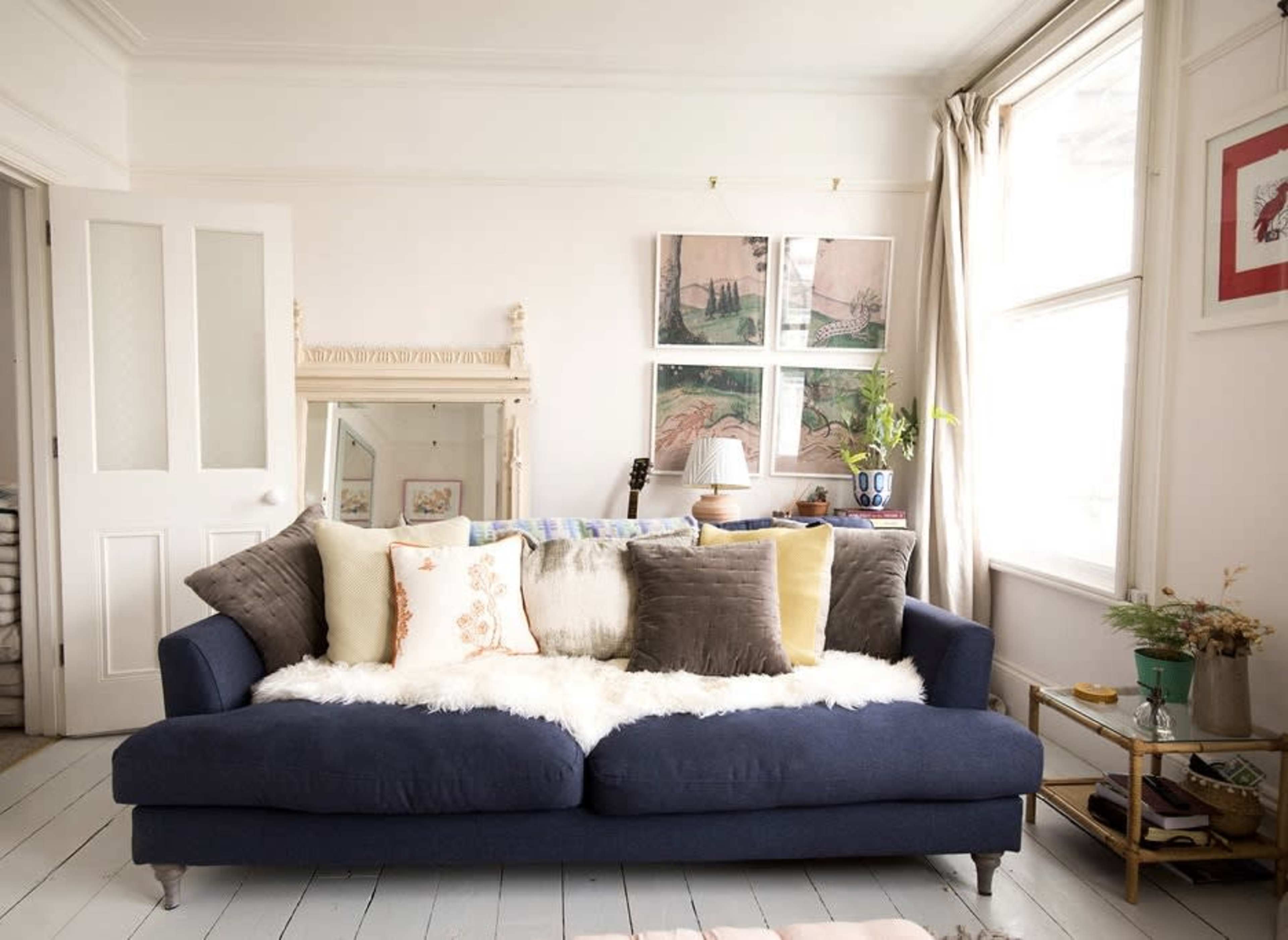 A blue sofa with several decorative pillows sits in a brightly lit room adorned with artwork and a mirror, while a small table holds a vase of flowers.