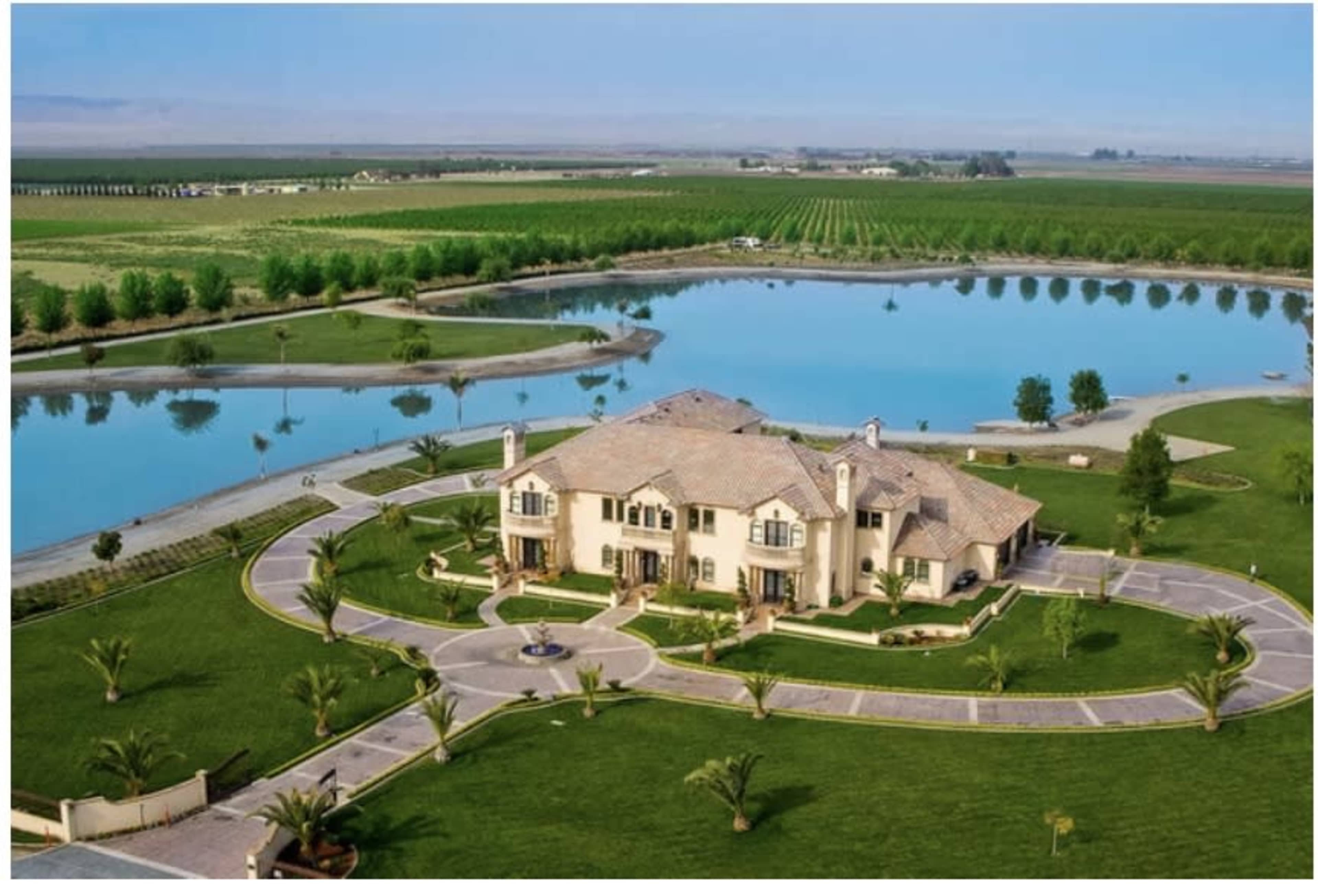 A large estate with a circular driveway, surrounded by well-manicured lawns, palm trees, and a reflective lake, set against a backdrop of agricultural fields.