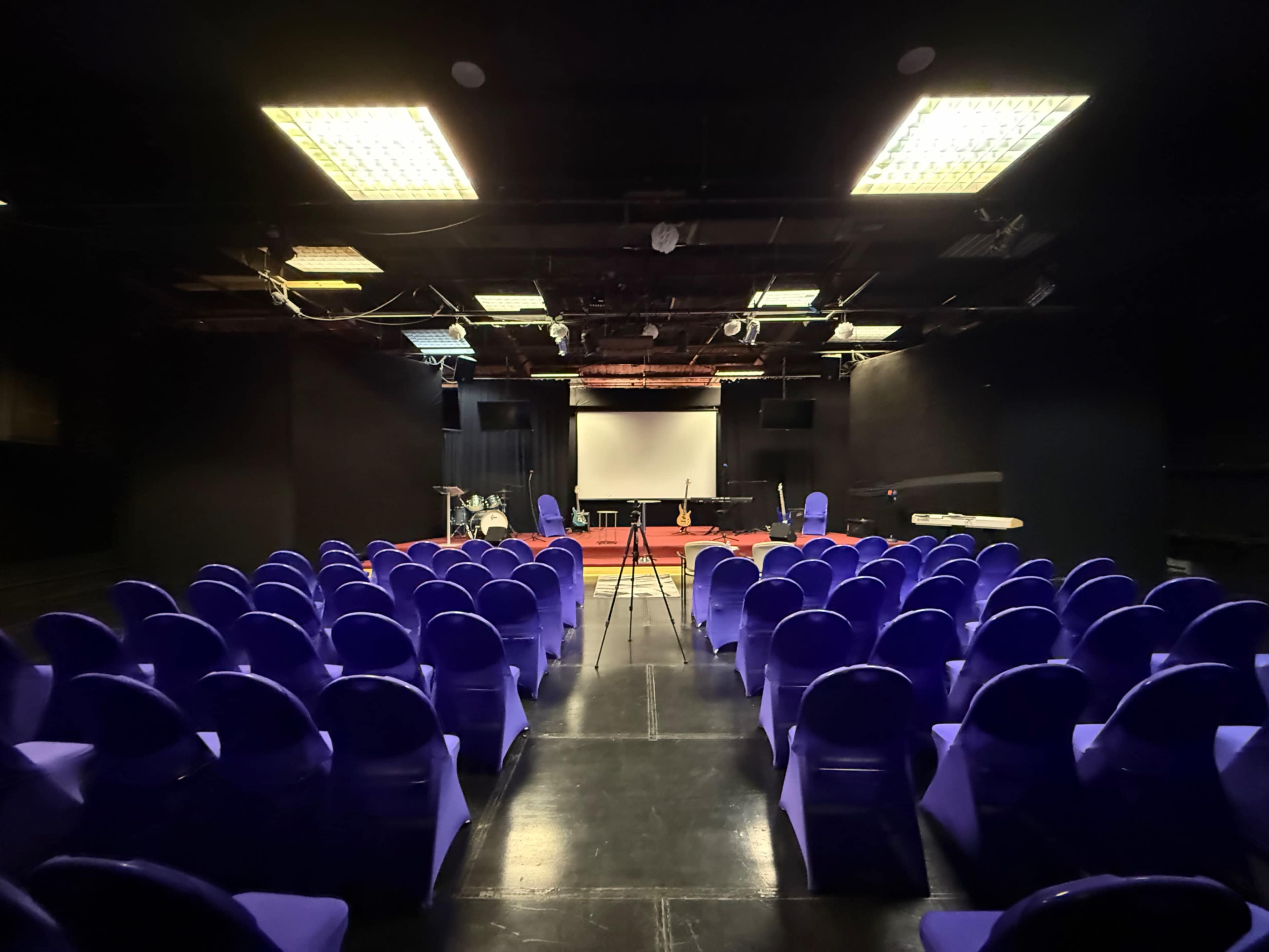 The image shows a dimly lit auditorium with rows of purple chairs facing a stage that features a blank projector screen and equipment set up for a presentation.