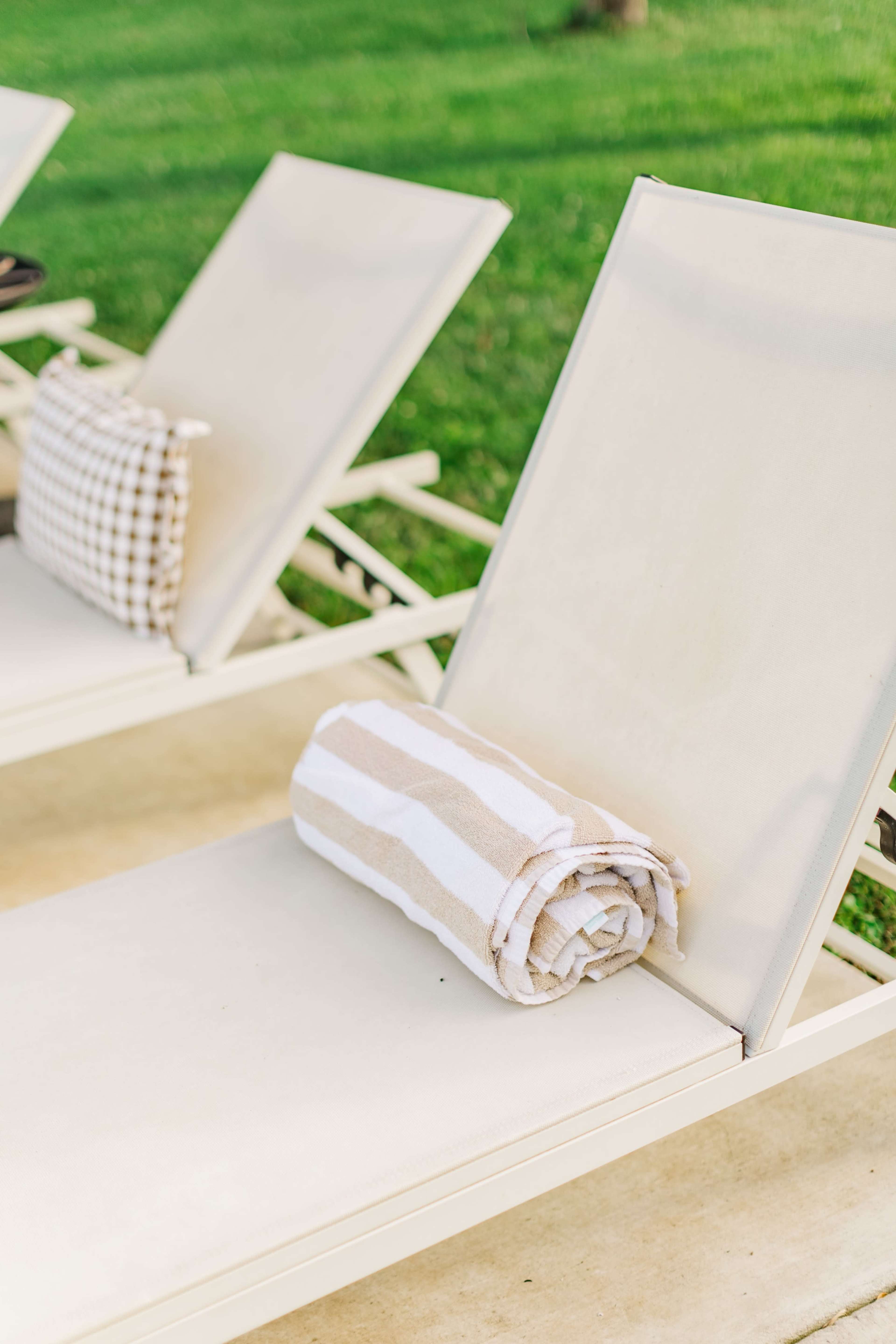 Two lounge chairs are positioned on a patio, each with a rolled towel resting on one of them, set against a green lawn.