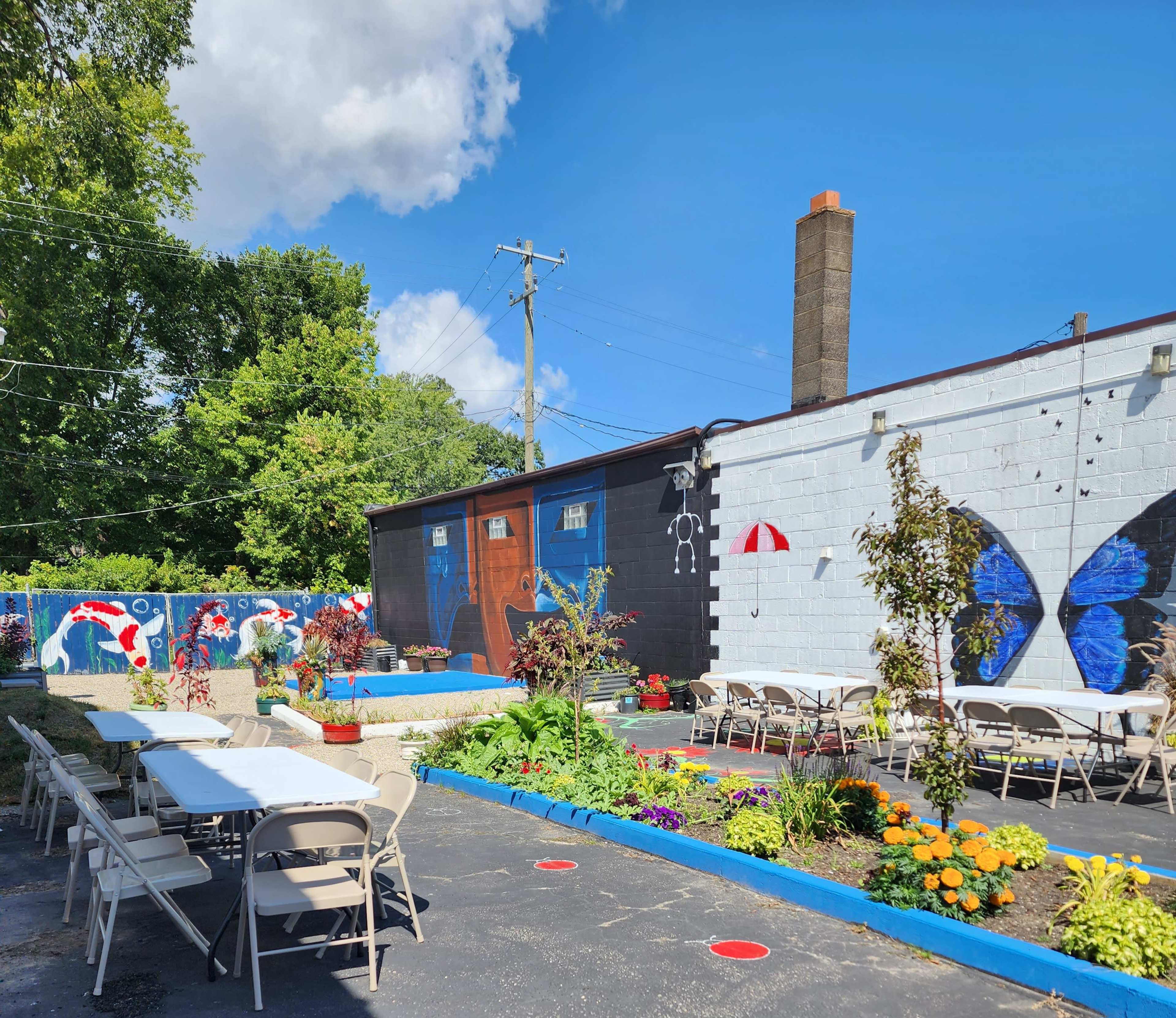 The image shows an outdoor space with tables set up in front of colorful murals on building walls, surrounded by greenery and flowers.