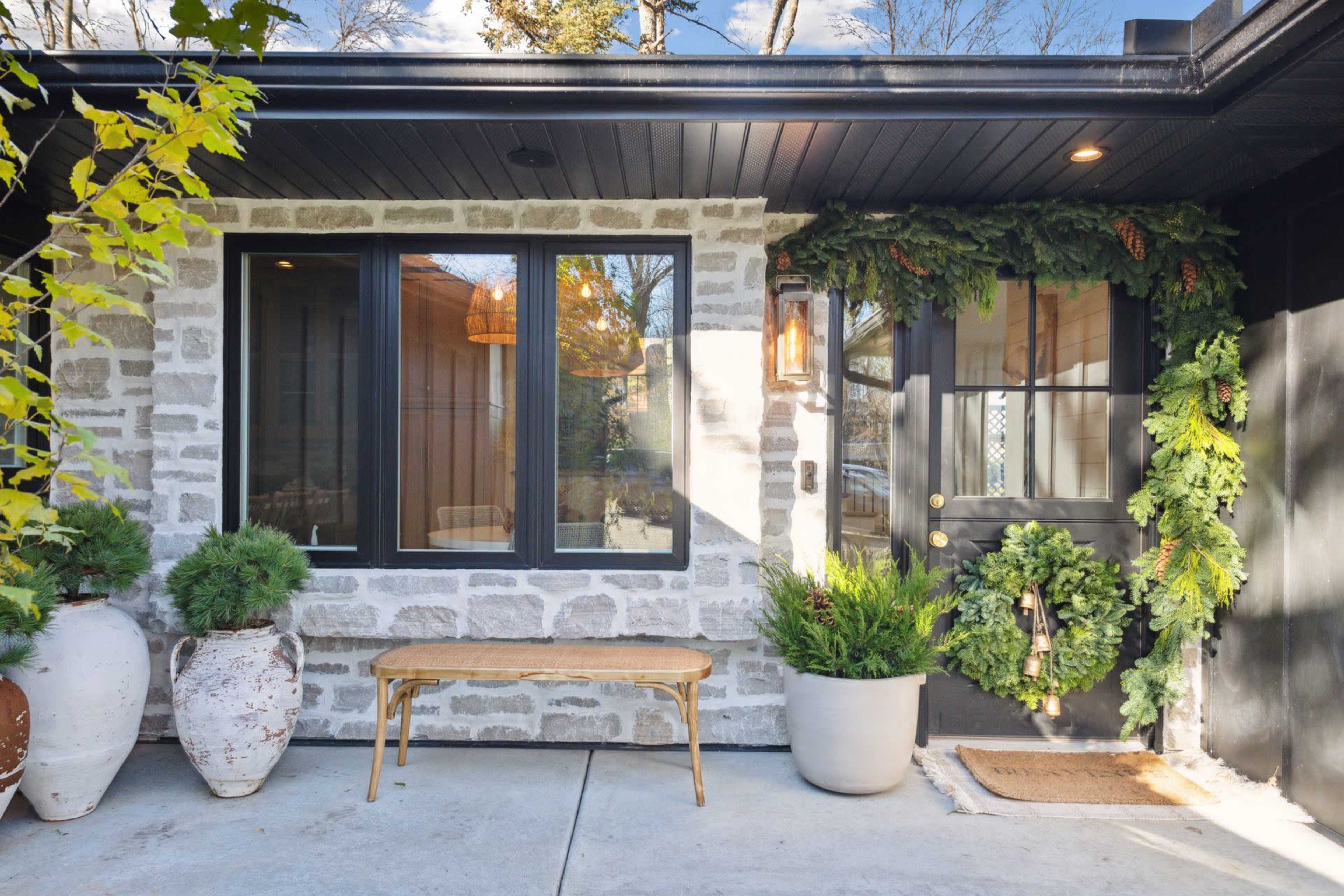 A front porch with a stone wall, large windows, a decorated door with a green wreath, a wooden bench, and potted plants.