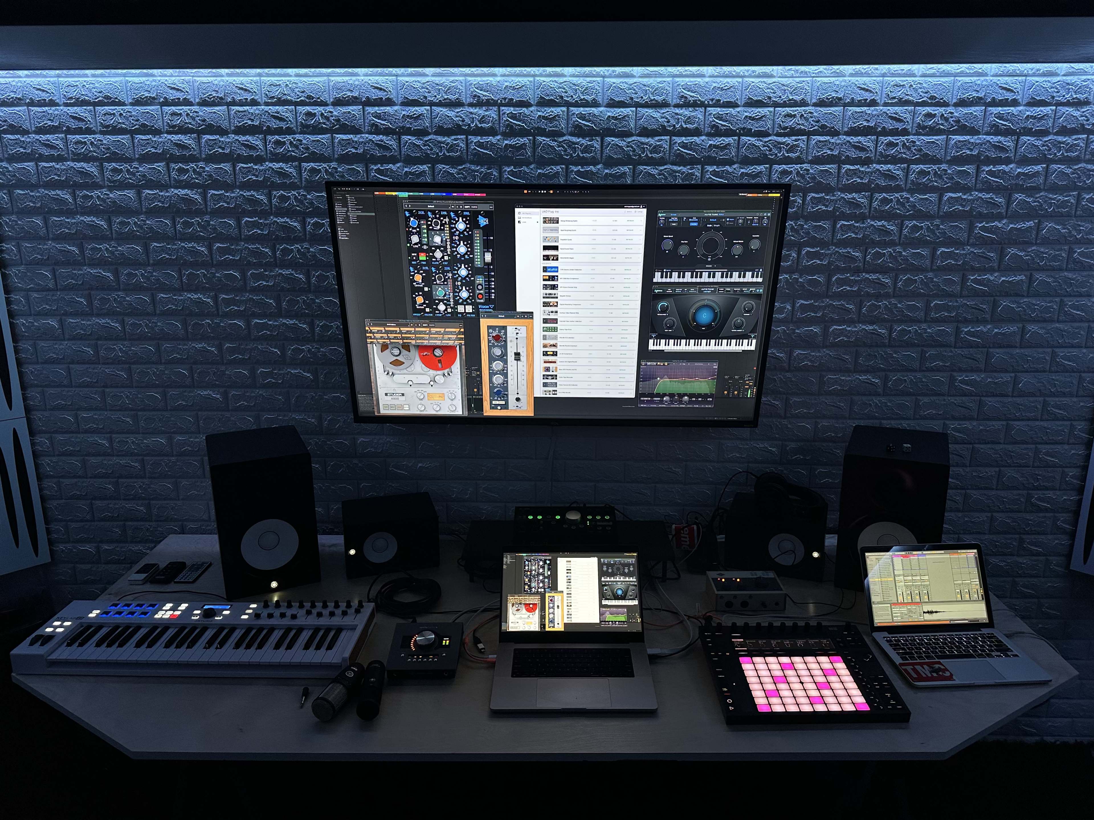 A music production setup features a large monitor displaying software, a laptop, and various electronic instruments arranged on a table with speakers and mixing equipment.