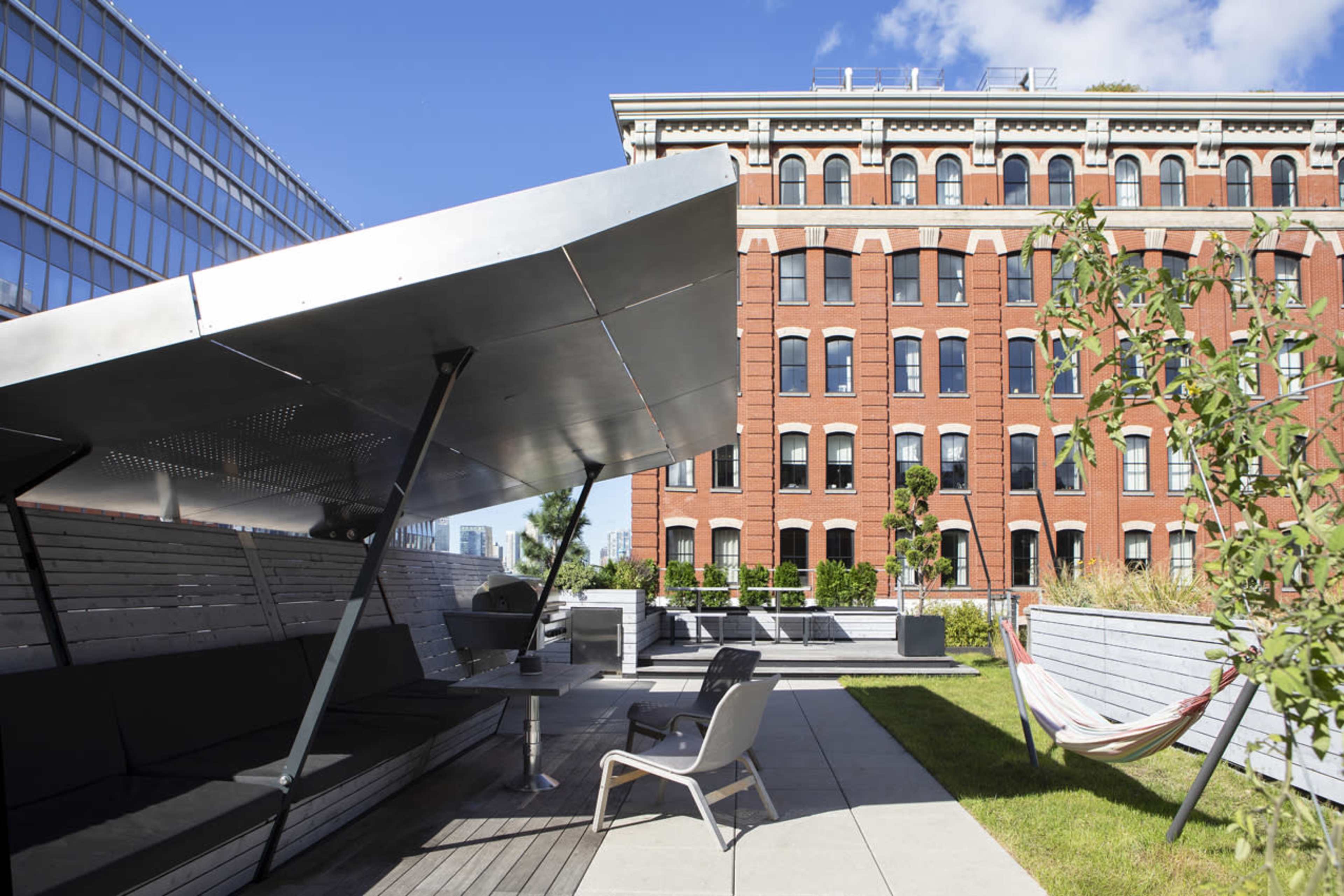 A modern rooftop space features angular metal seating under a large shade canopy, adjacent to a brick building with numerous windows.