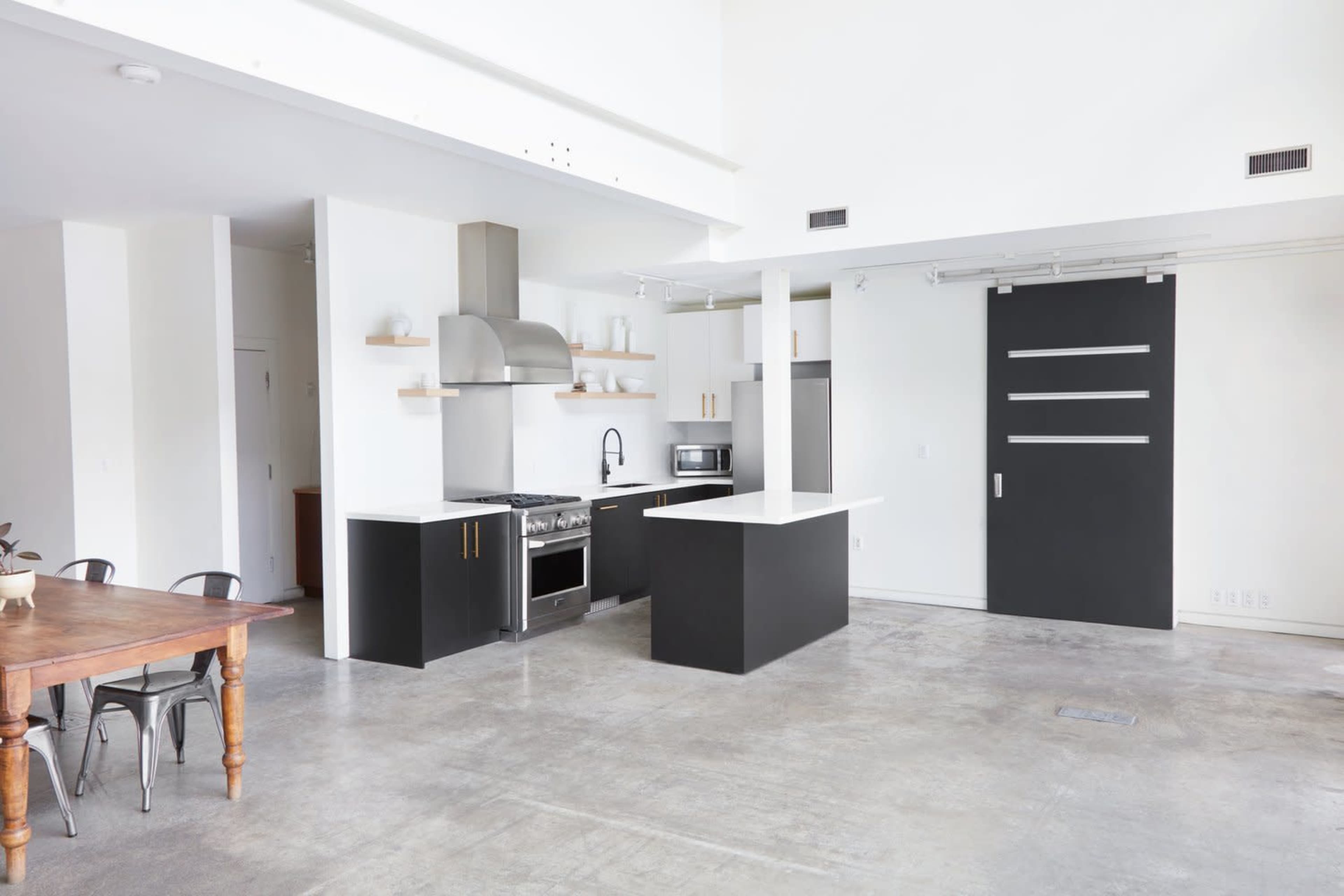 A modern kitchen with a black island, stainless steel appliances, and a concrete floor, adjacent to a dining area with a wooden table and metal chairs.