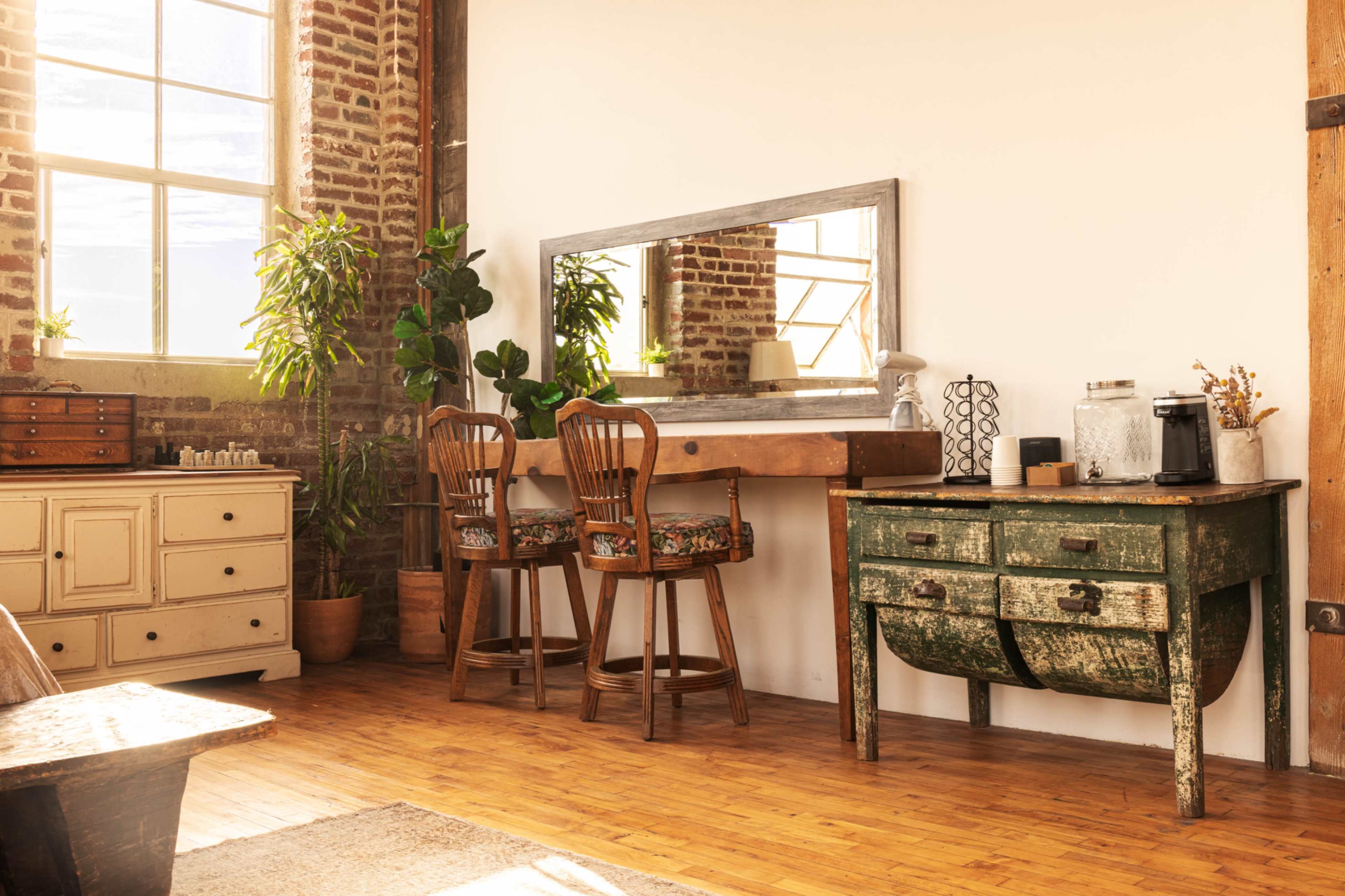 A bright, rustic room with wooden flooring, a large mirror above a wooden table, and two chairs, surrounded by plants and various furniture pieces.