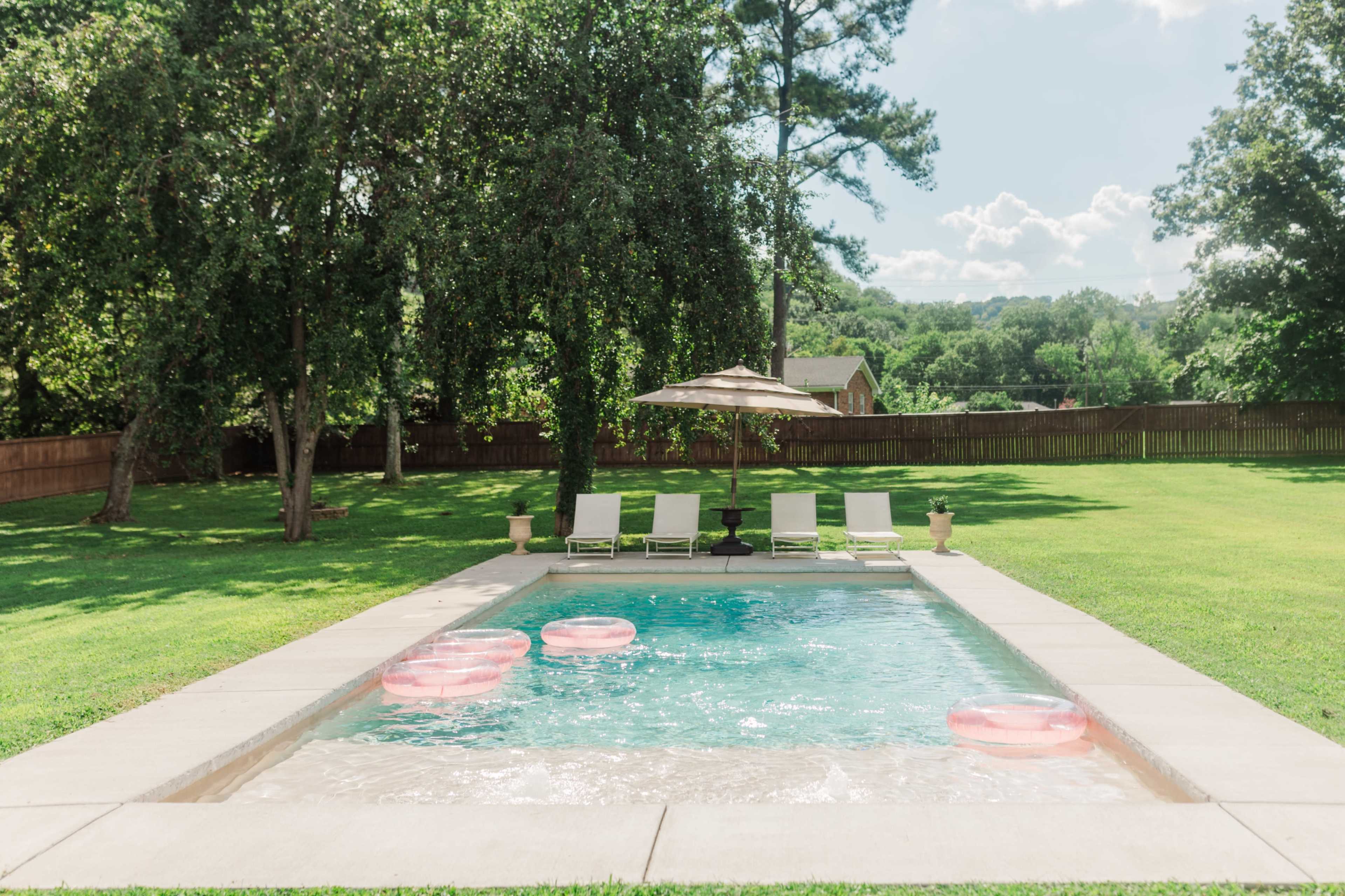 A swimming pool surrounded by green grass features pink inflatable floats and lounge chairs under a sun umbrella.