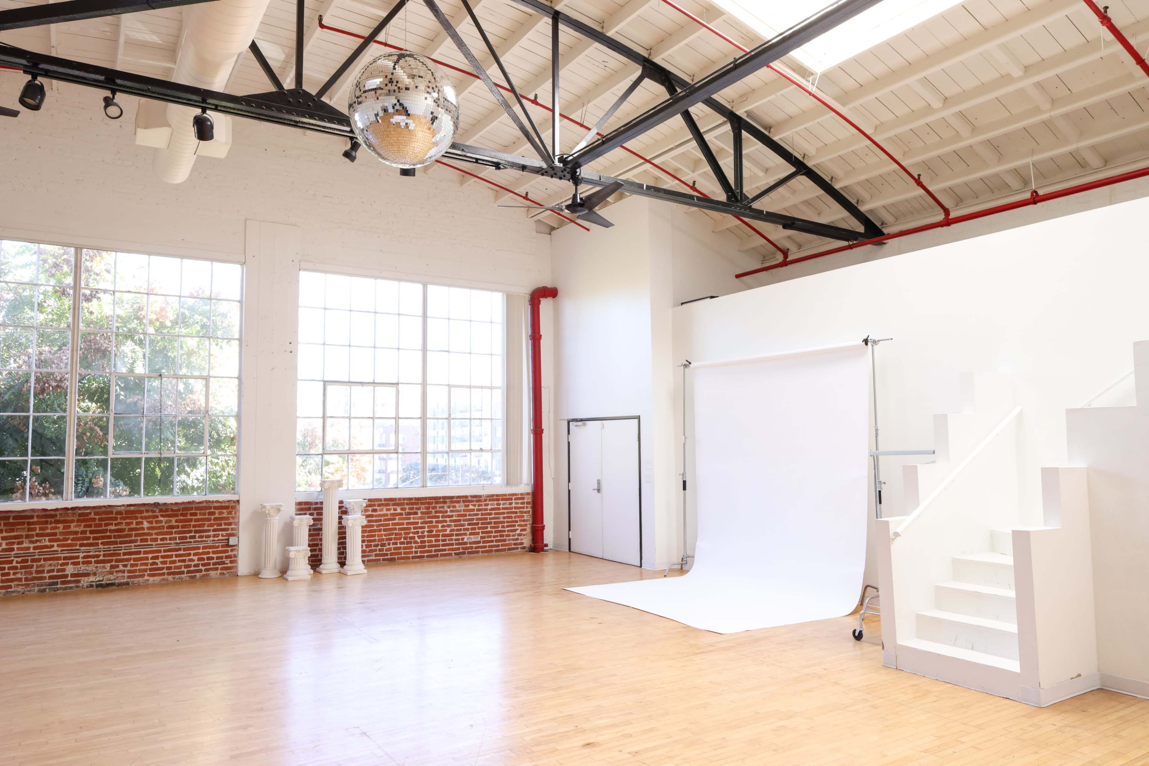 A bright, spacious studio with large windows, wooden flooring, a white backdrop for photography, and a staircase leading to a raised area, all under a disco ball hanging from the ceiling.
