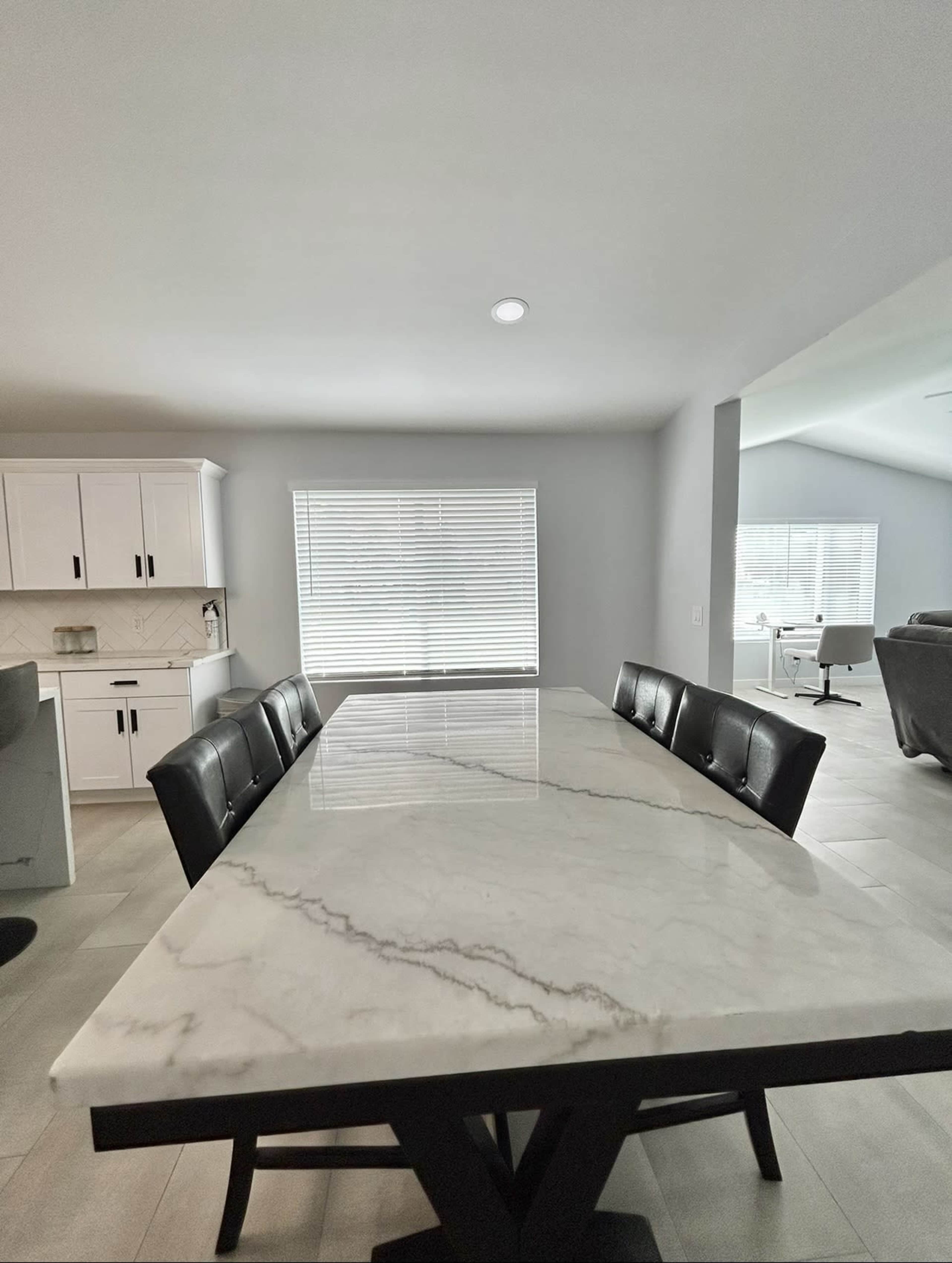 A modern dining area features a large marble-top table surrounded by black upholstered chairs, with natural light coming through a window.