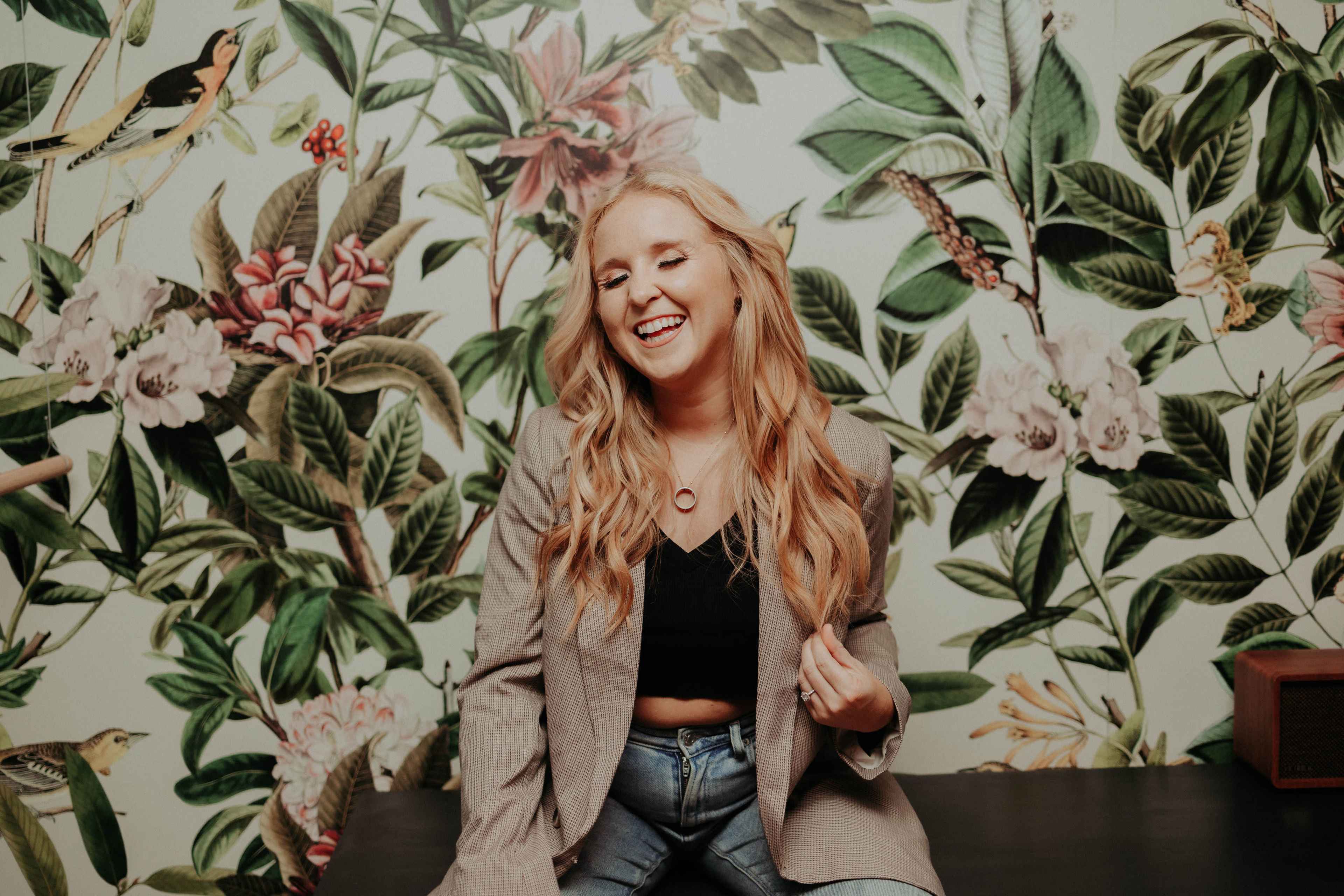 A woman with long blonde hair sits in front of a floral wallpaper, wearing a blazer and jeans, smiling as she poses.