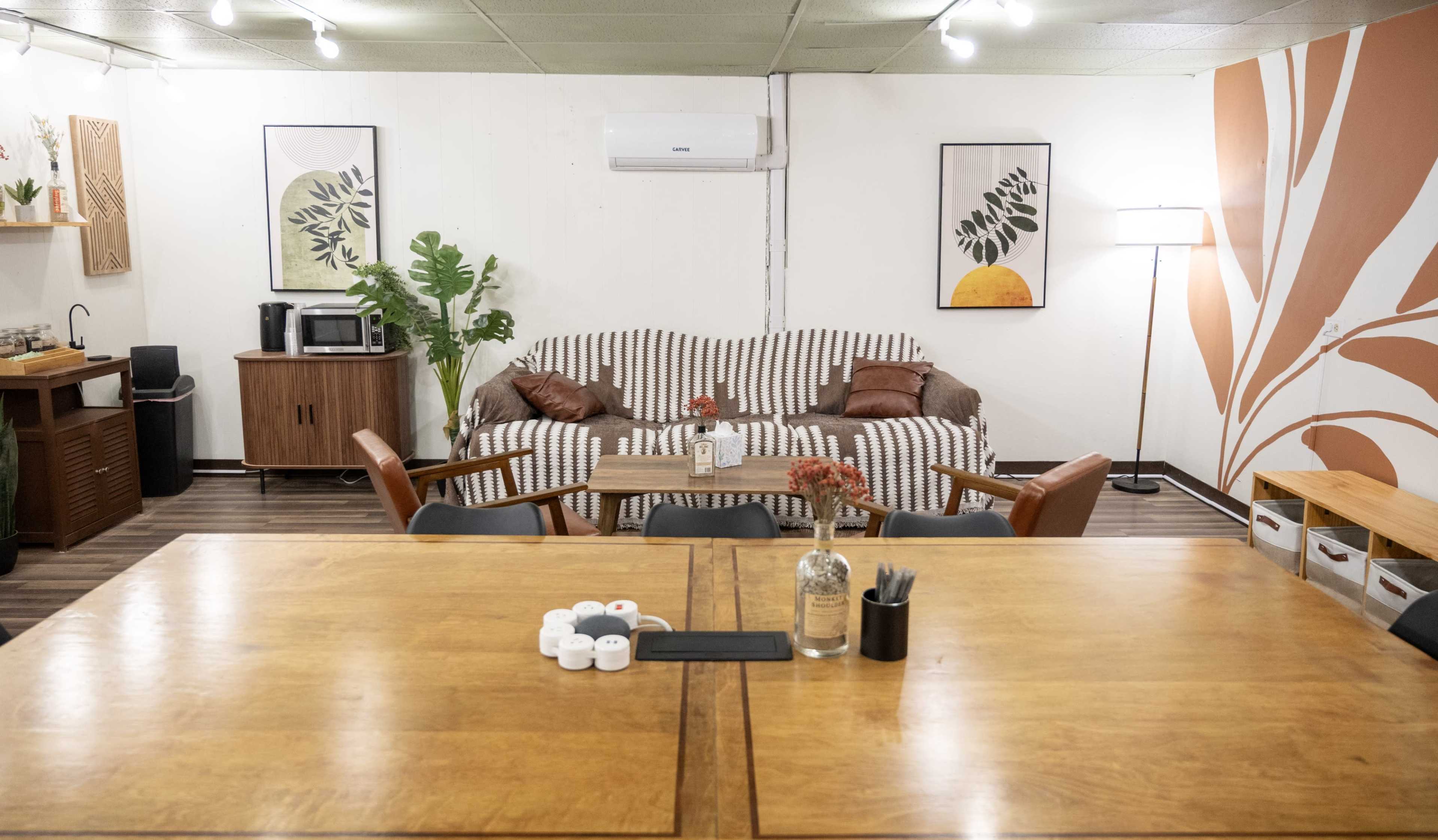 The image shows a cozy, well-decorated room with a brown sofa, a wooden table, and decorative plants, illuminated by overhead lights and a floor lamp.