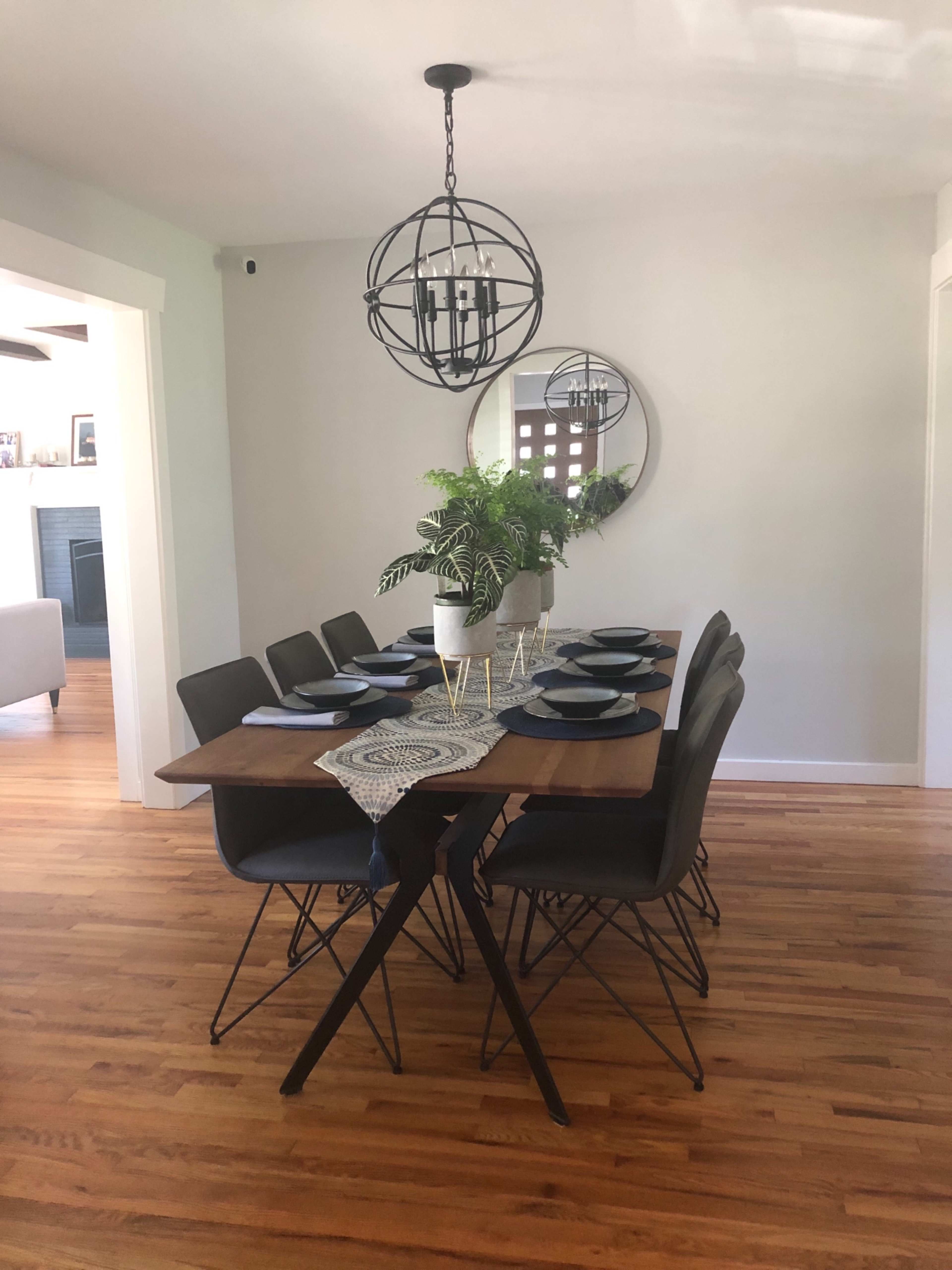 A wooden dining table with eight black chairs is set with blue plates and a decorative runner, beneath a round chandelier and a circular mirror on the wall.