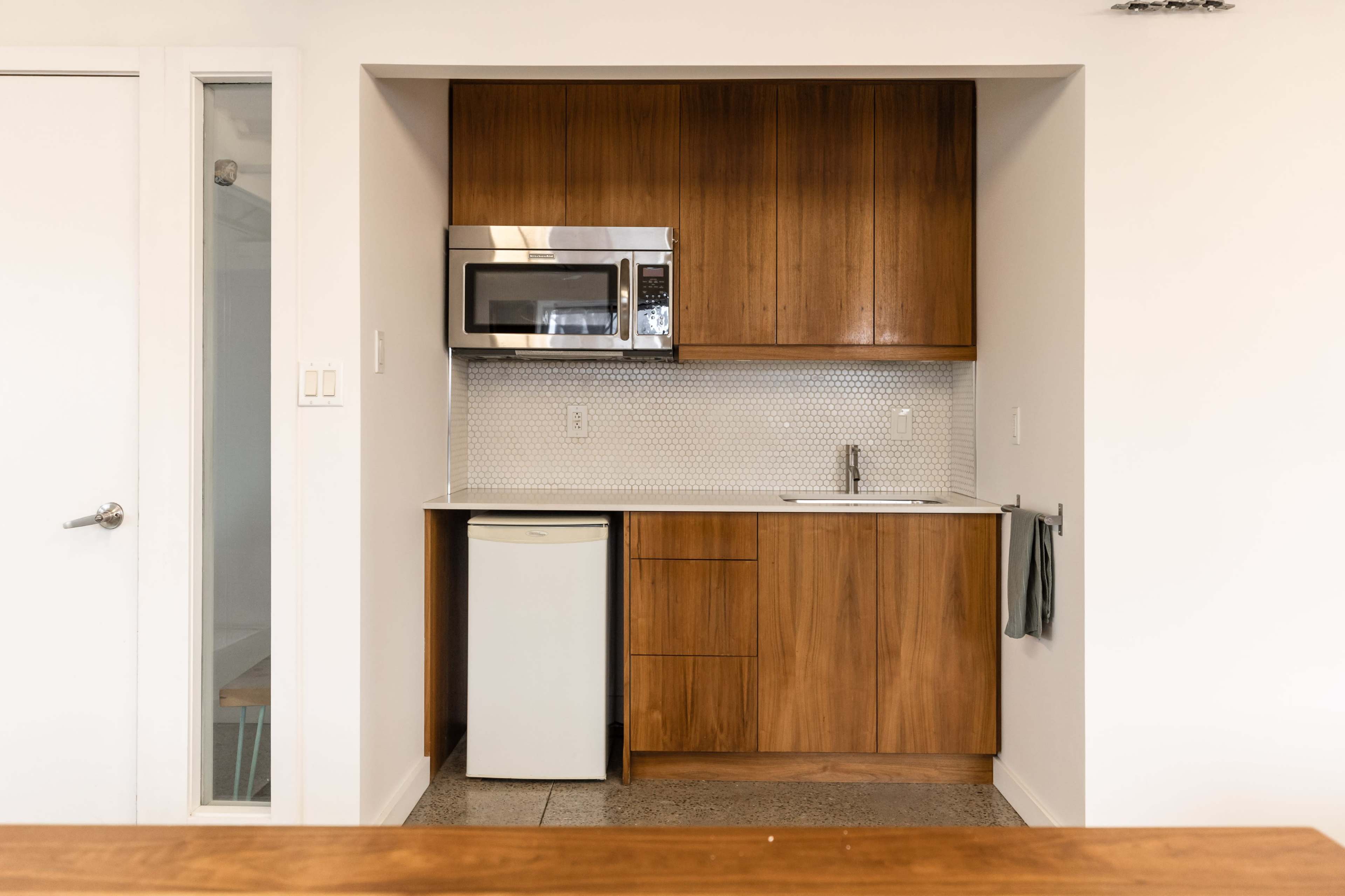 The image shows a modern kitchen area featuring wooden cabinetry, a microwave, a small refrigerator, and a sink with a backsplash of white tiles.
