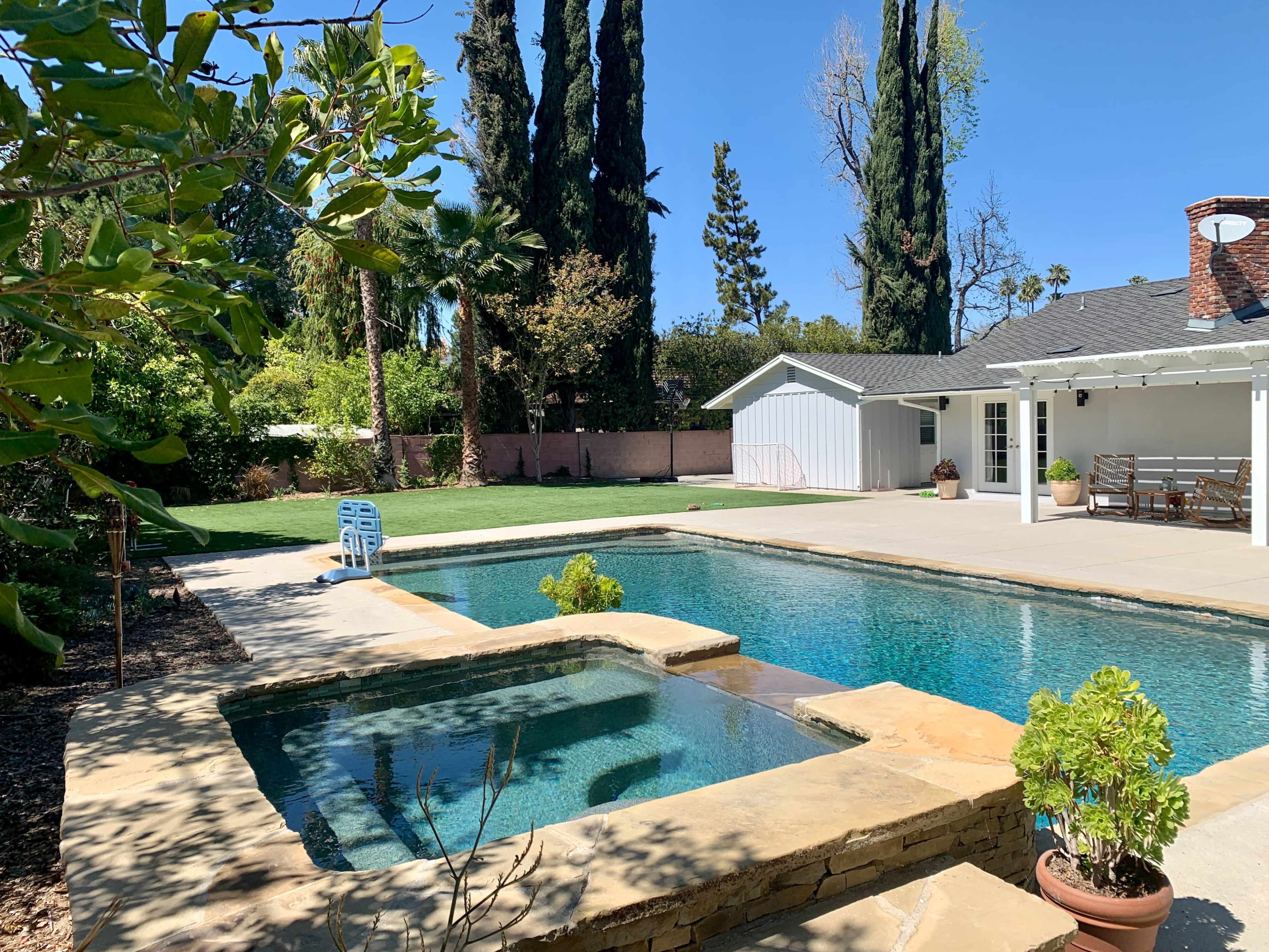 A swimming pool with a spa, surrounded by greenery and a single-story house in the background.