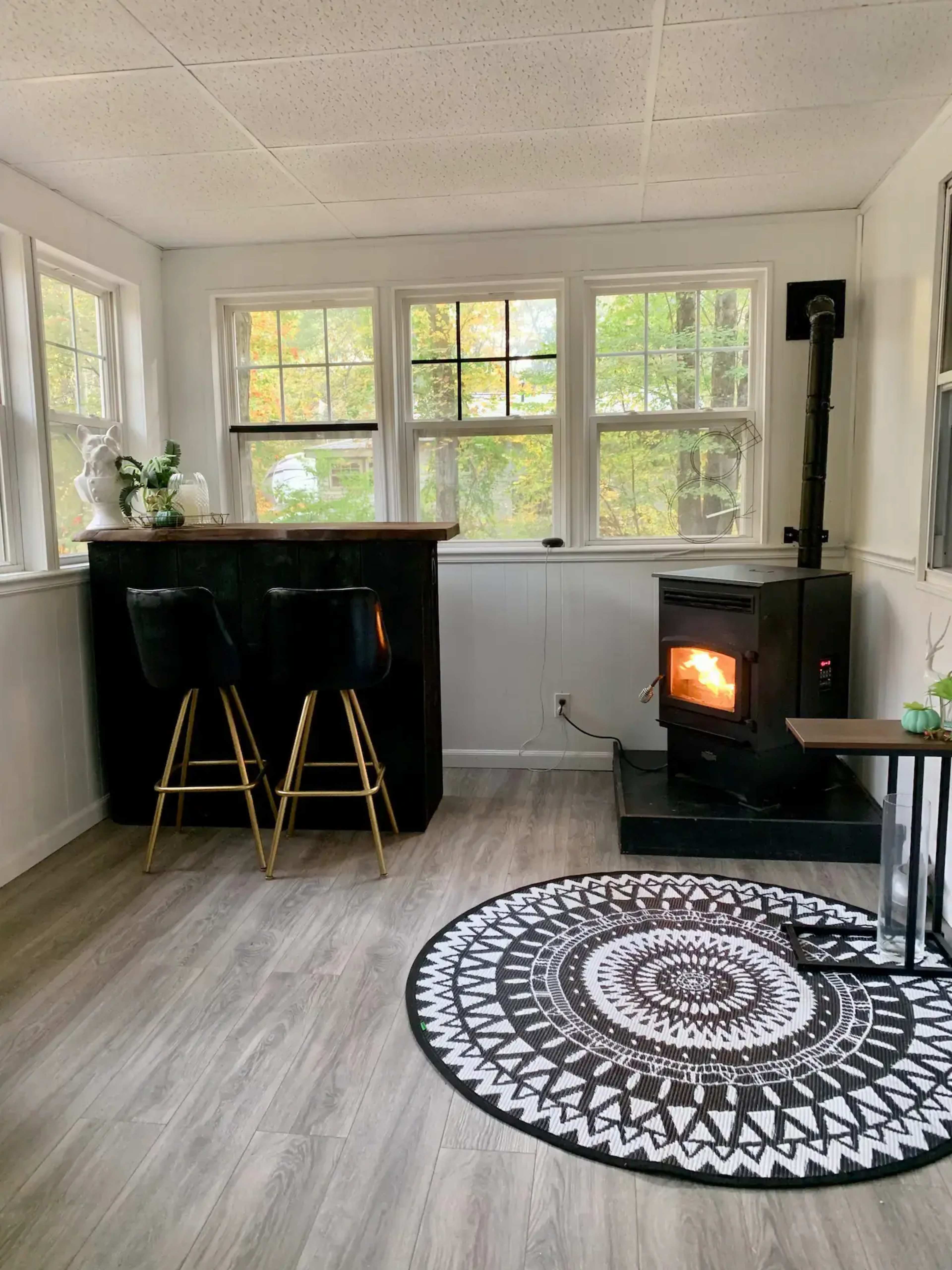 A cozy sitting area features a black stove with a visible flame, two bar stools at a counter, and a patterned rug on a wood floor, surrounded by windows showcasing greenery outside.
