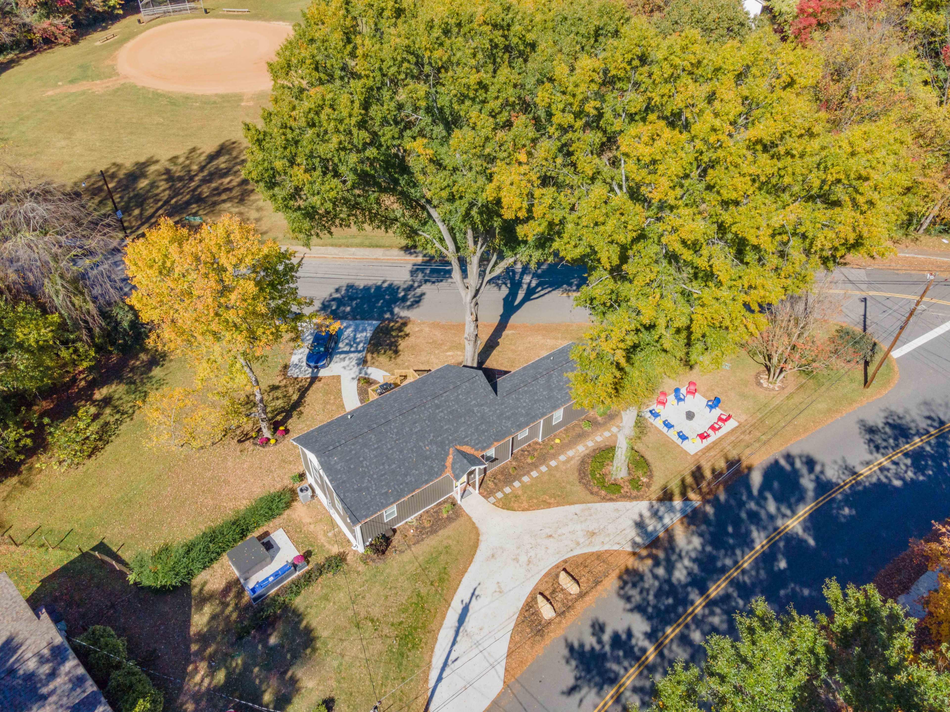 The image shows a house situated at the corner of a street near a baseball field, surrounded by trees and a landscaped yard.