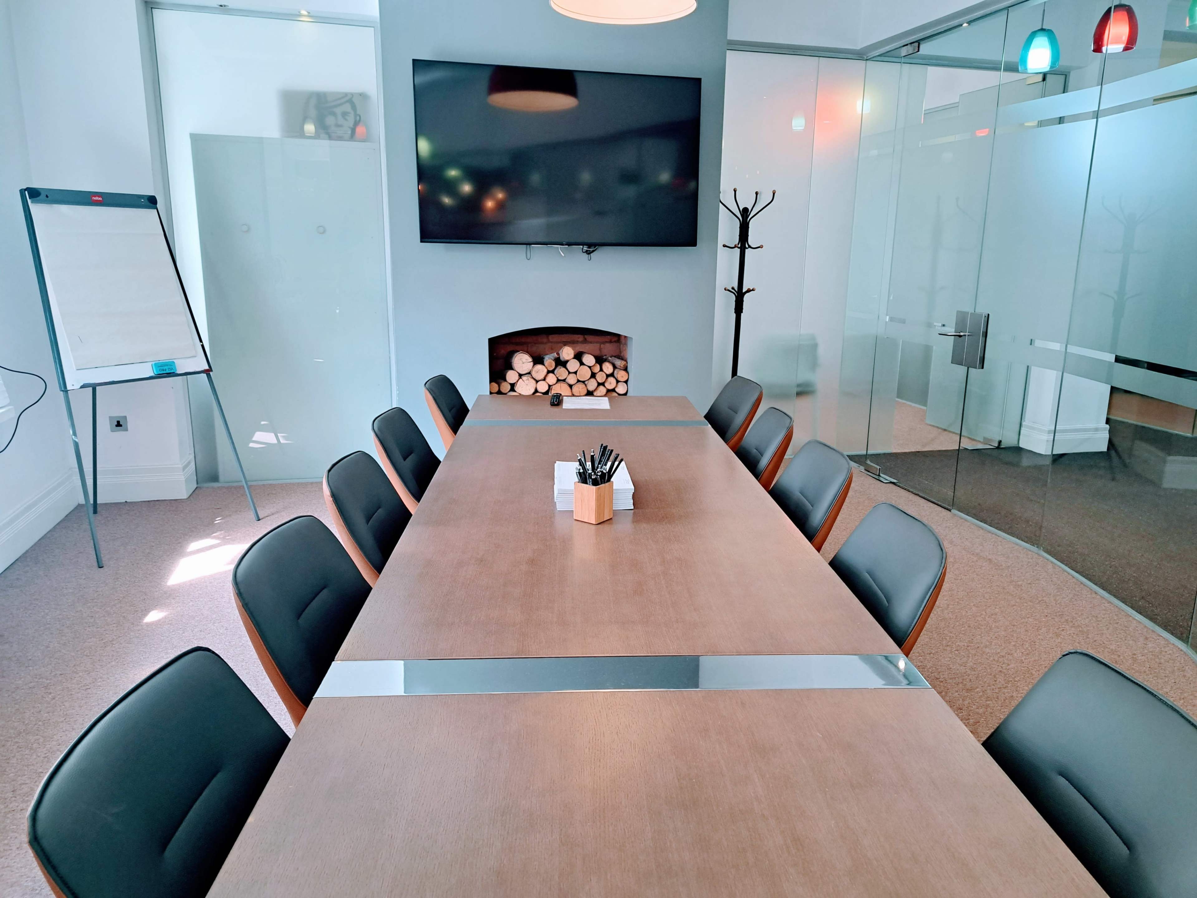 A modern conference room features a long wooden table with black chairs, a television mounted on the wall, and a fireplace displaying logs.