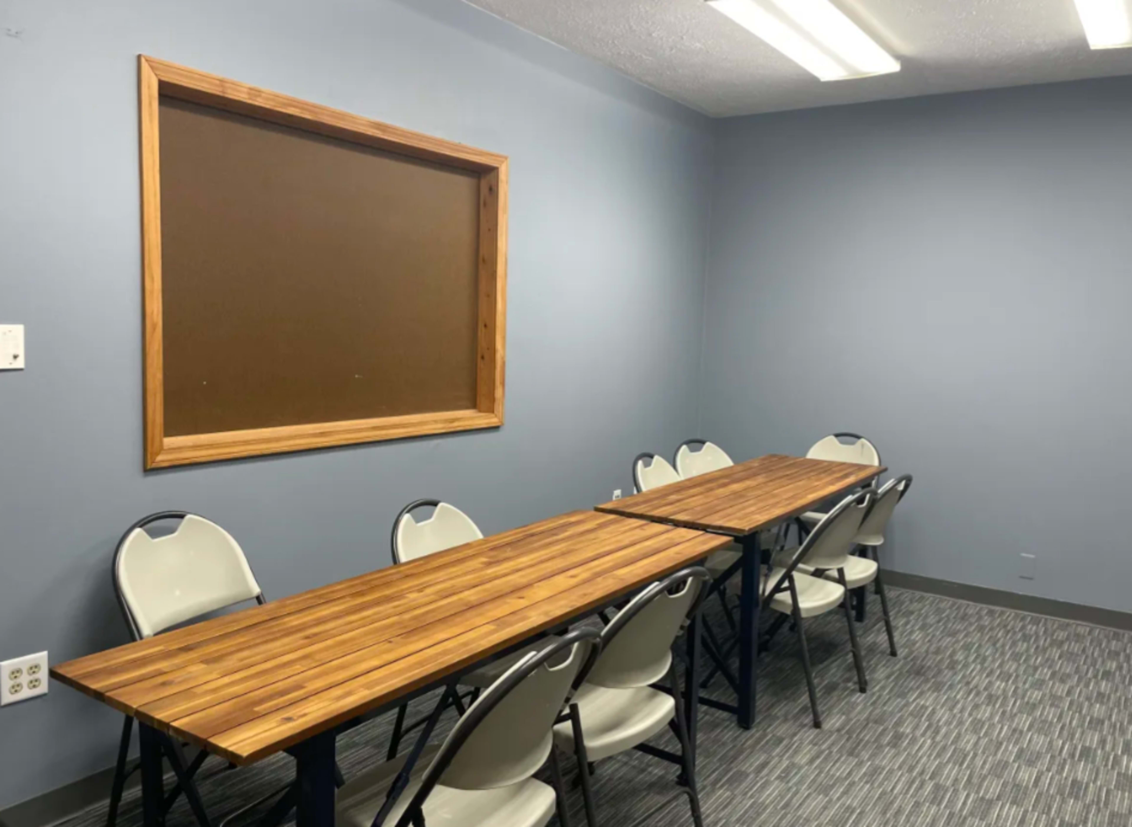 The image shows a small meeting room featuring a long wooden table and several plastic chairs against a blue wall with a bulletin board.