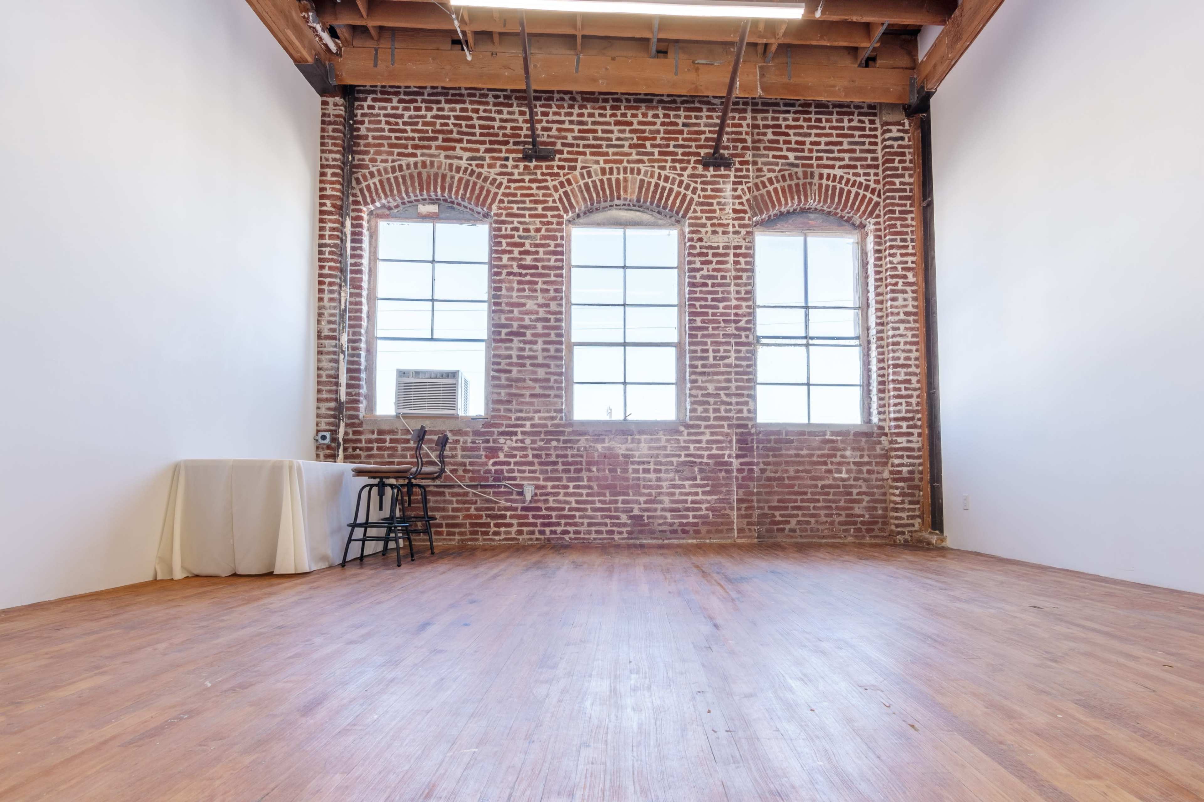 A spacious room with exposed brick walls, large windows, and wooden flooring.