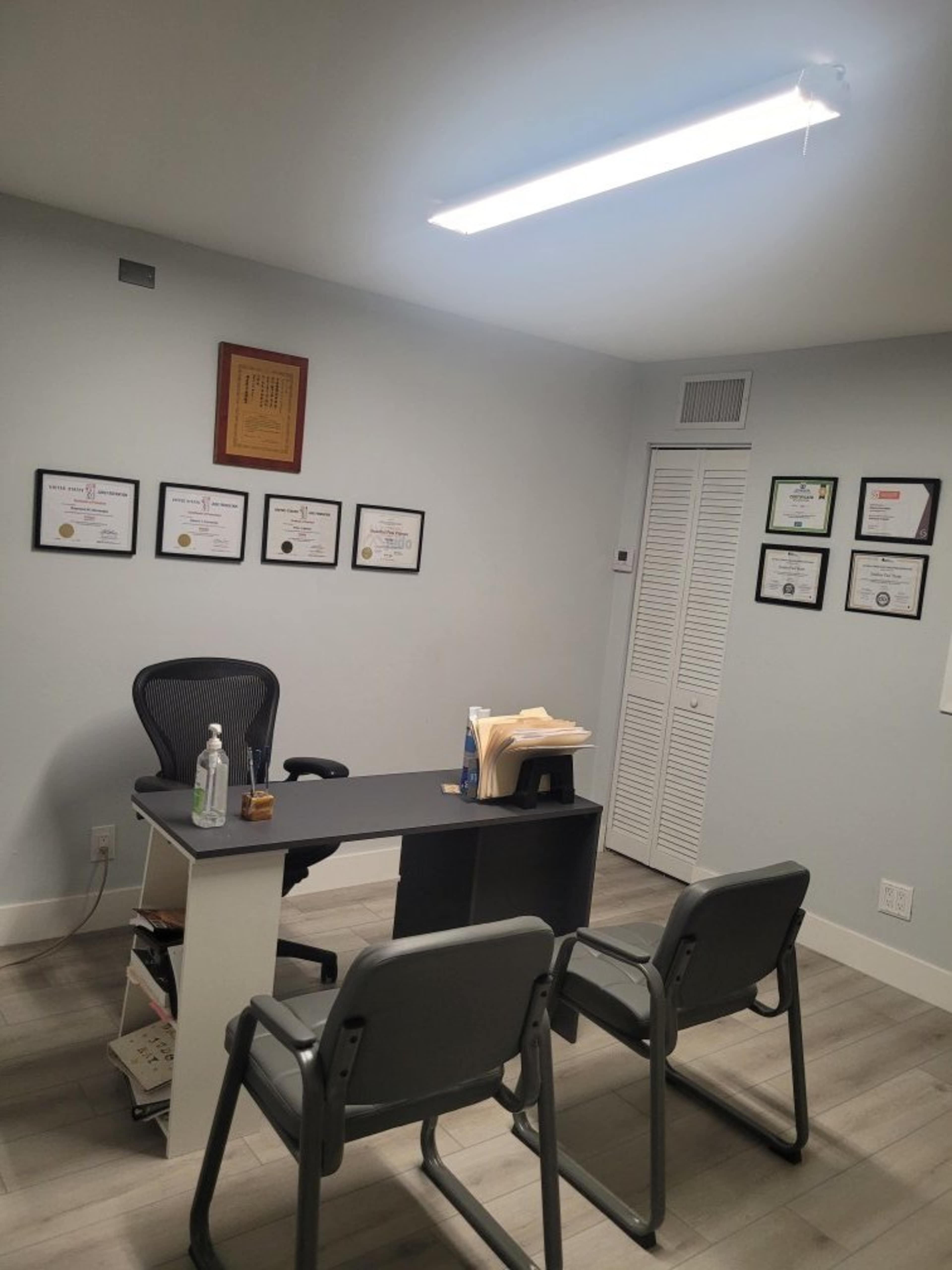 The image shows a small office with a desk, two chairs in front, a wall adorned with framed certificates, and a closet door in the back.
