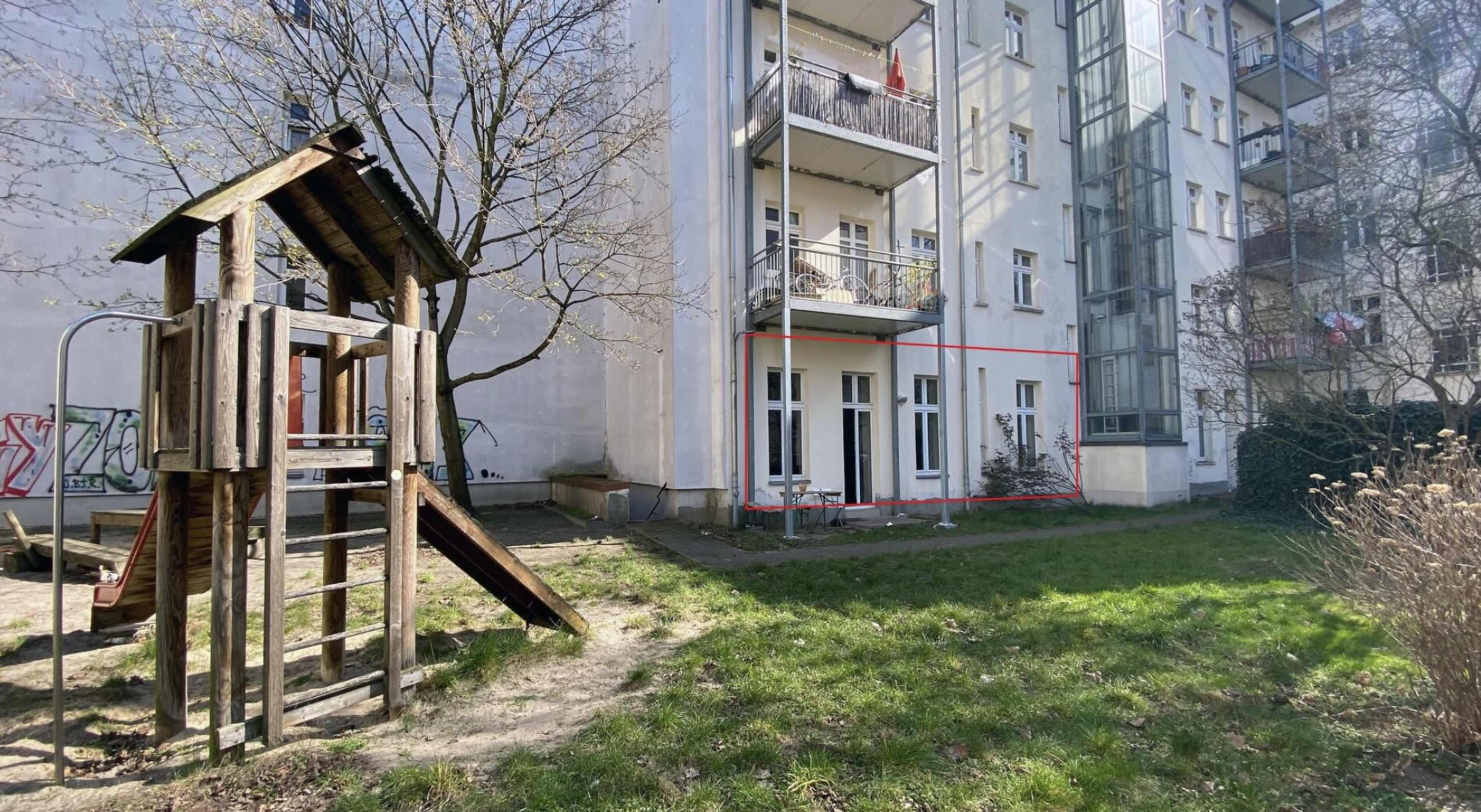 A playground with a wooden play structure beside a building that has balconies and a garden area.