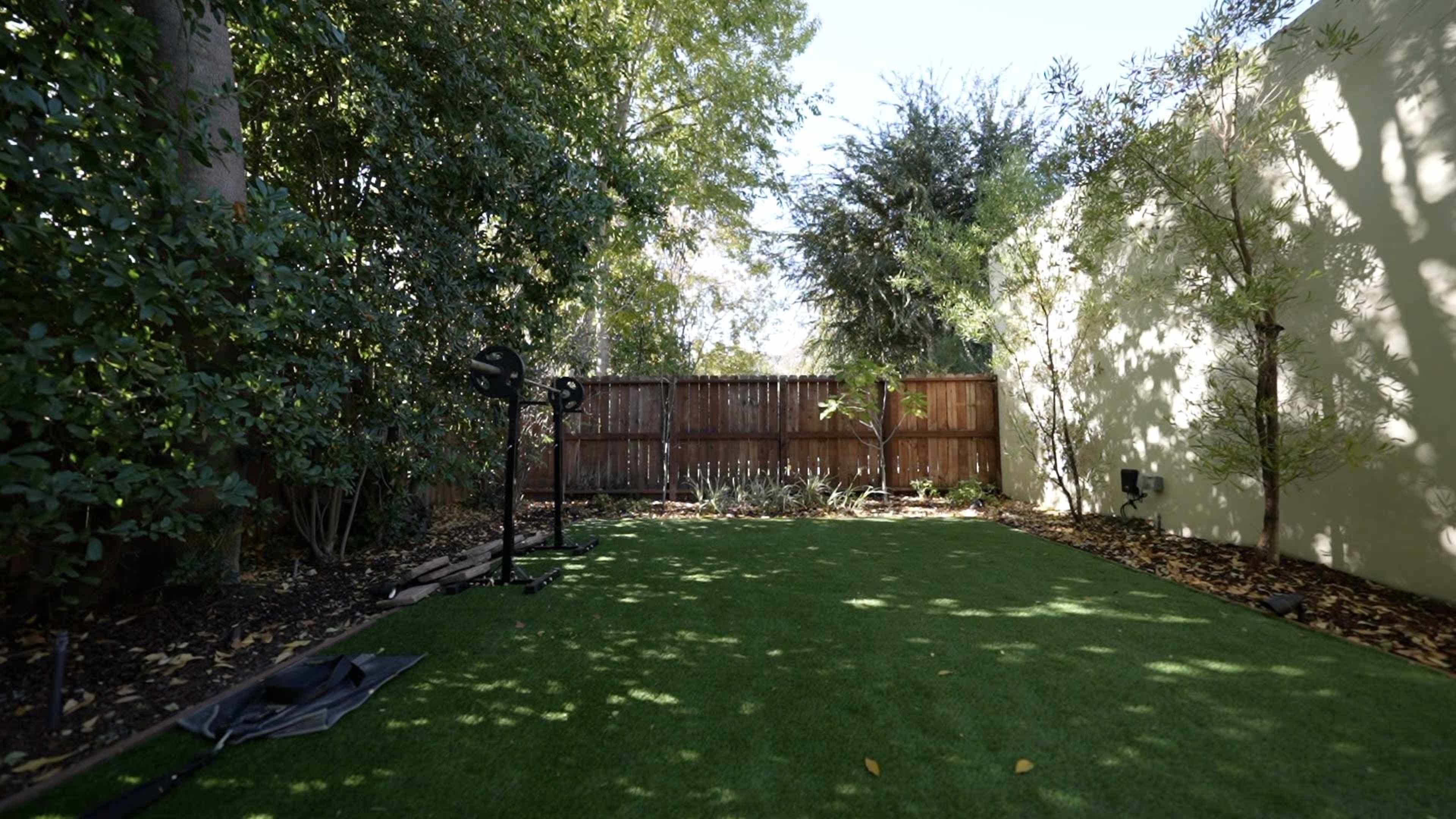 A fenced backyard with artificial grass, surrounded by trees and shrubs, featuring a basketball hoop in one corner.