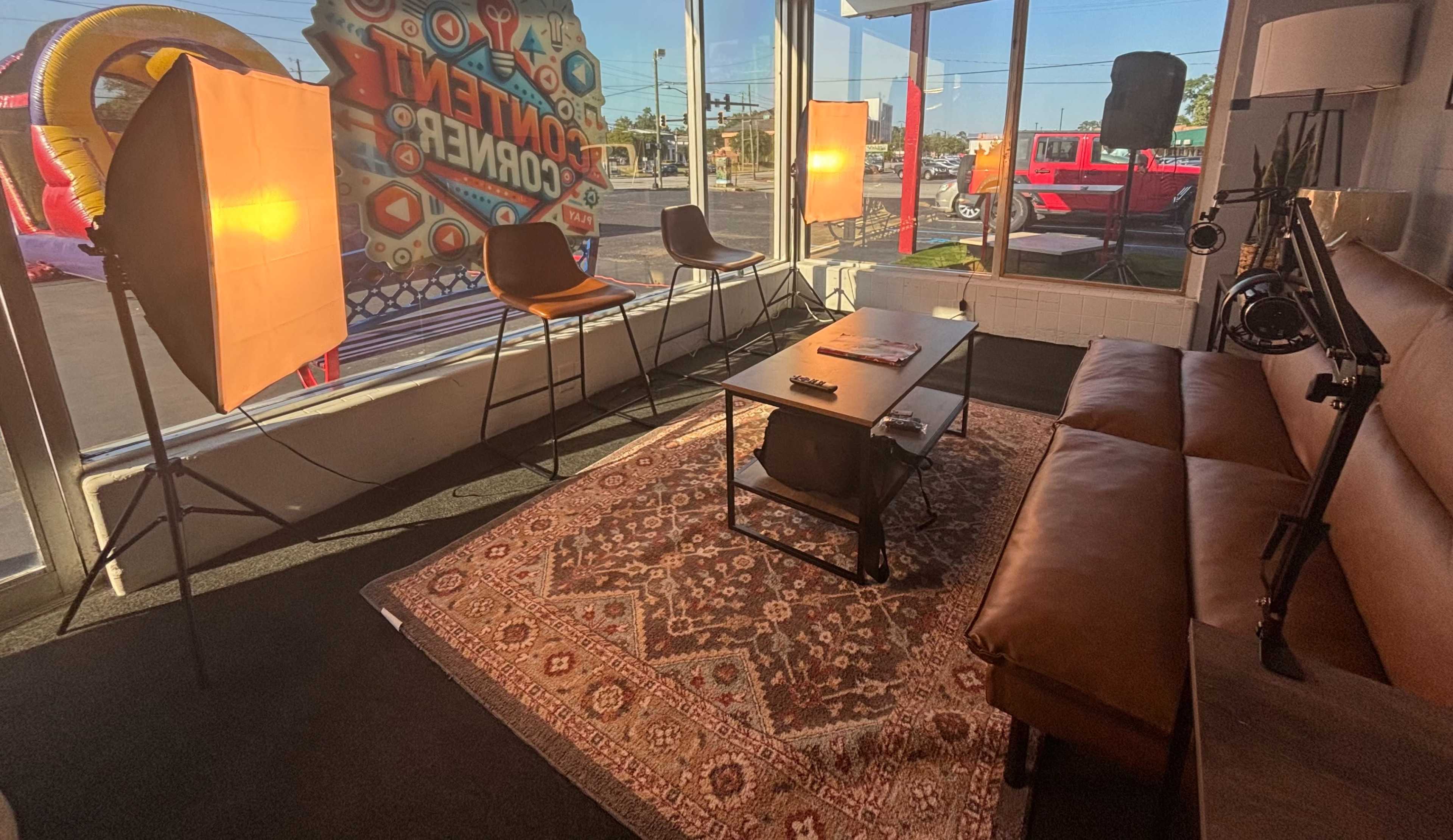 The image shows a well-lit corner of a casual interior space with a sofa, coffee table, and decorative rug, featuring large windows and studio lights.