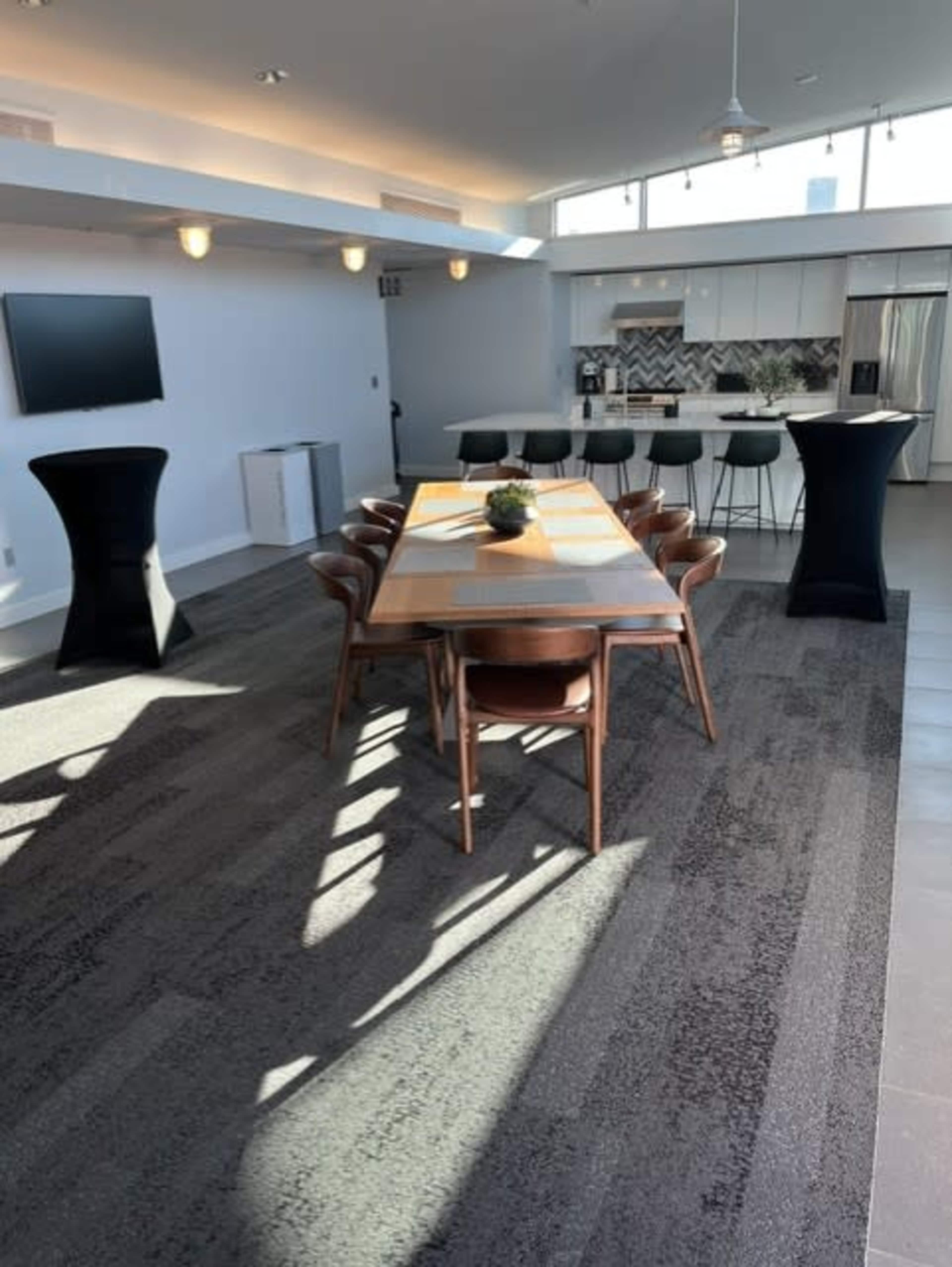 The image shows a modern dining area with a long wooden table, black chairs, and a kitchen in the background, featuring a countertop and appliances.