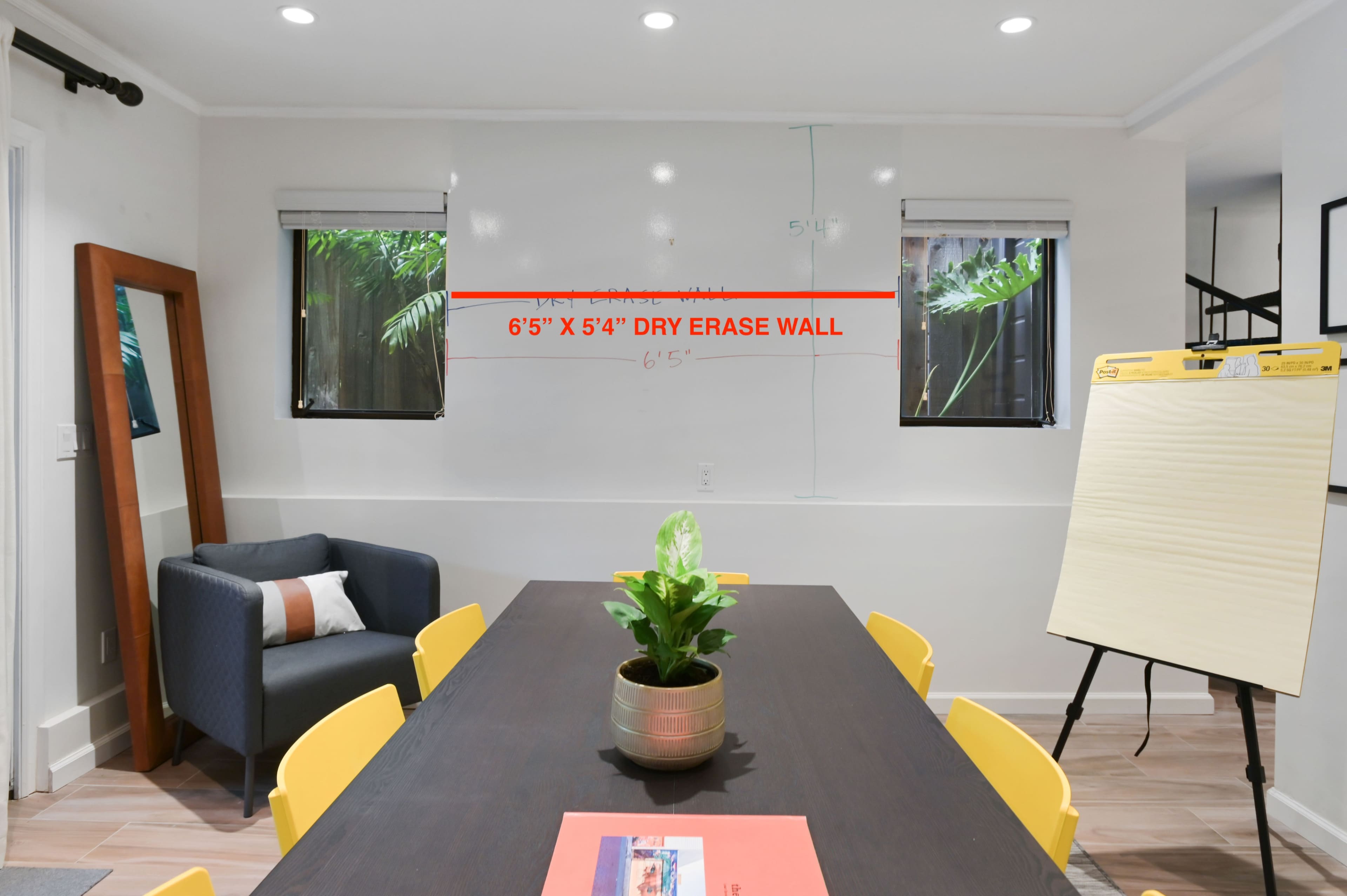 A conference room features a large dry erase wall with a mirror and windows, a long dining table surrounded by yellow chairs, and a potted plant as the centerpiece.