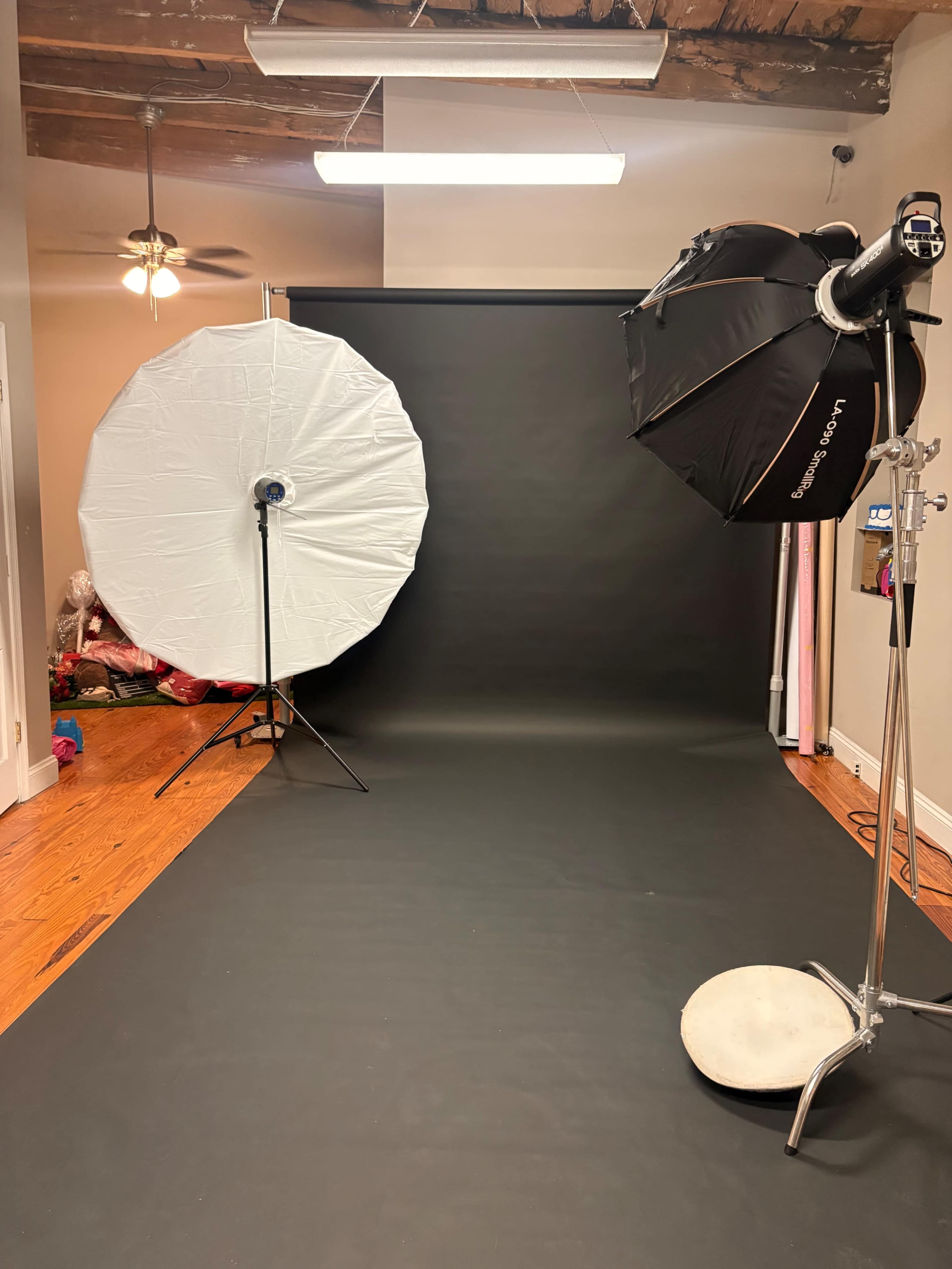 The image shows a photography setup with two light sources, one with a white umbrella and the other with a black softbox, both positioned in front of a dark backdrop.