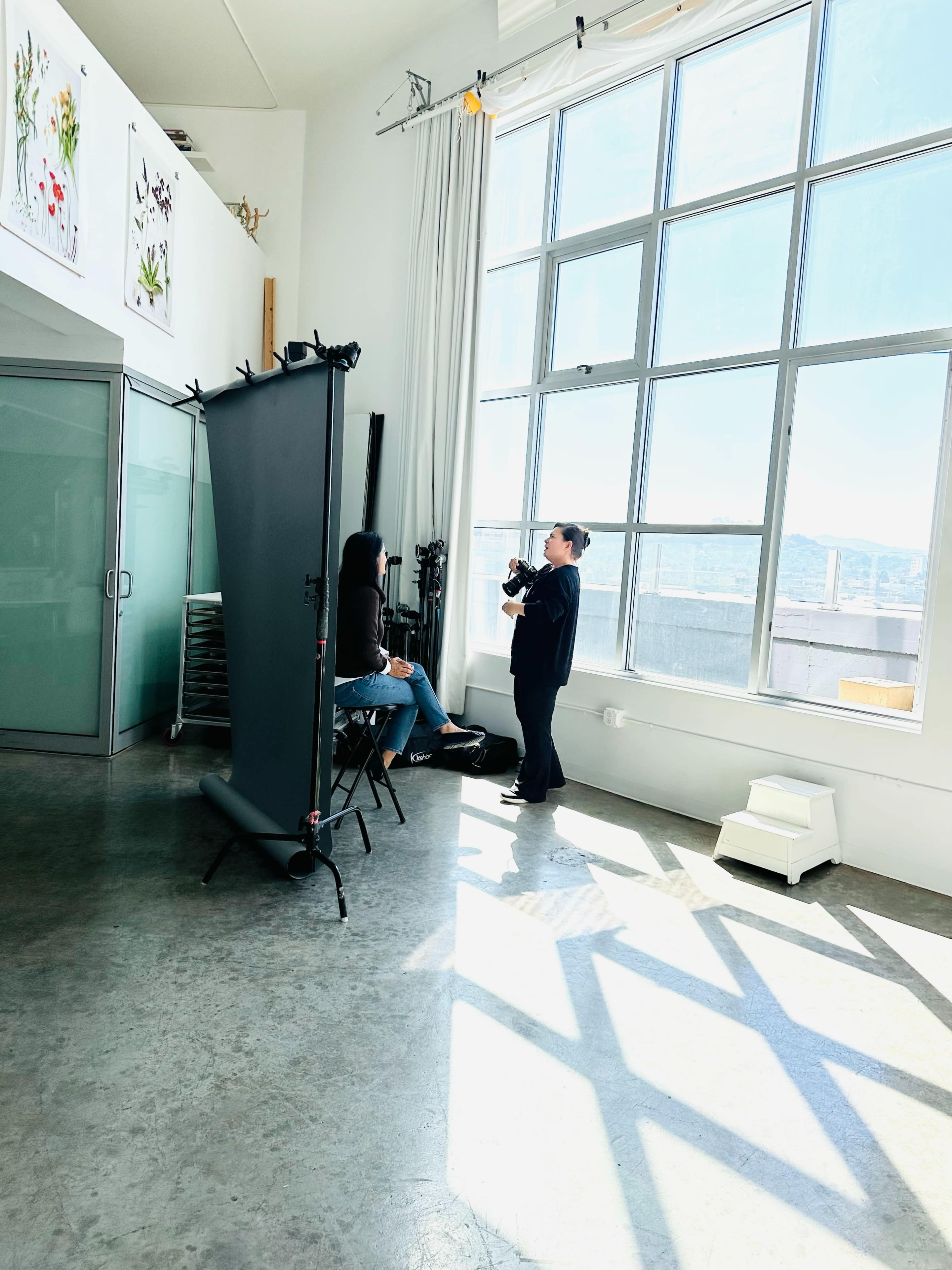 A photographer captures images of a model in a well-lit studio with large windows and a gray backdrop.