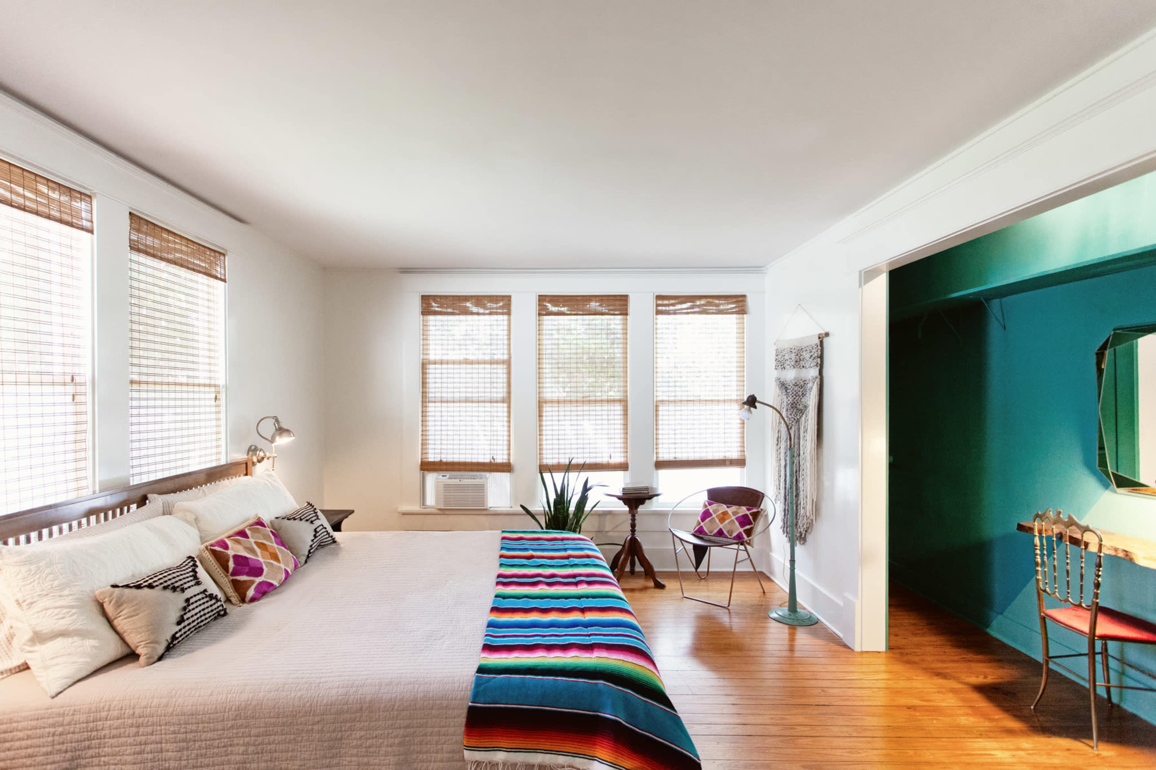The bedroom features a large bed with colorful bedding, natural light streaming through multiple window blinds, and a small desk area in an adjacent space.