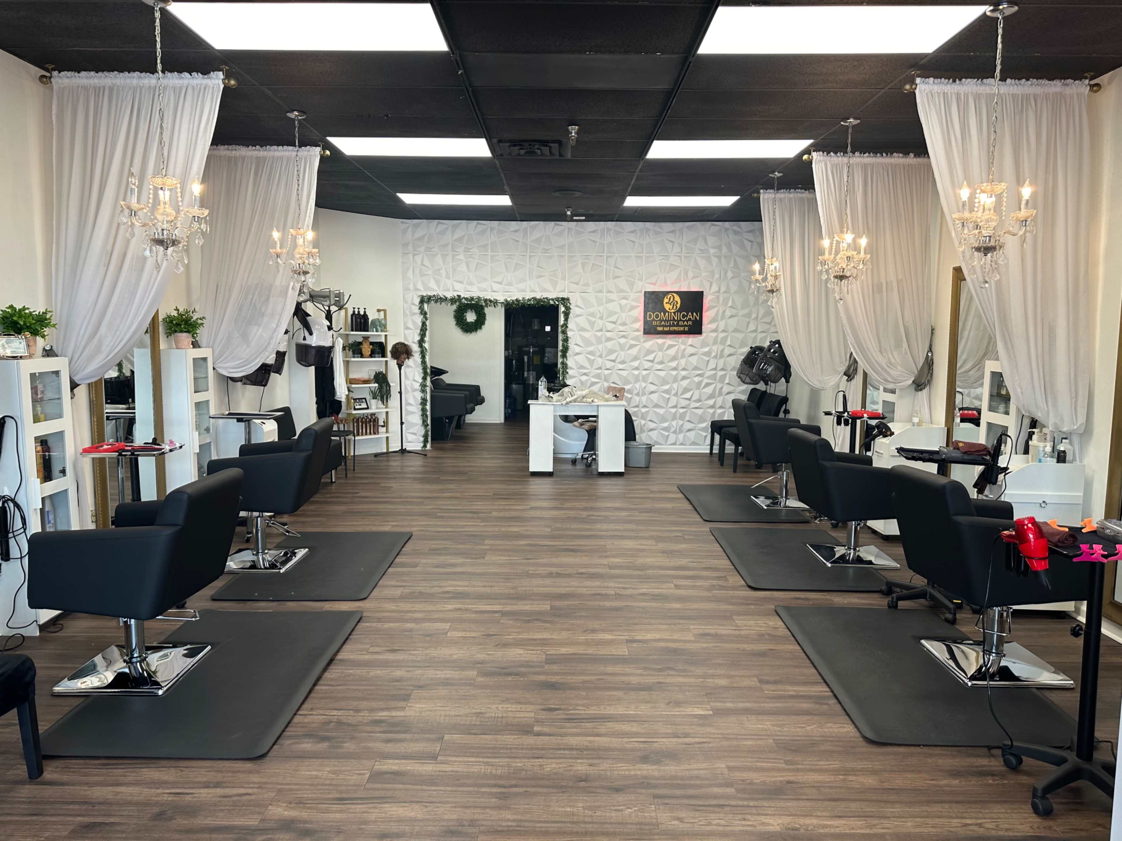 The image shows a spacious hair salon with several styling stations featuring black chairs and white shelving, decorated with sheer curtains and chandeliers.
