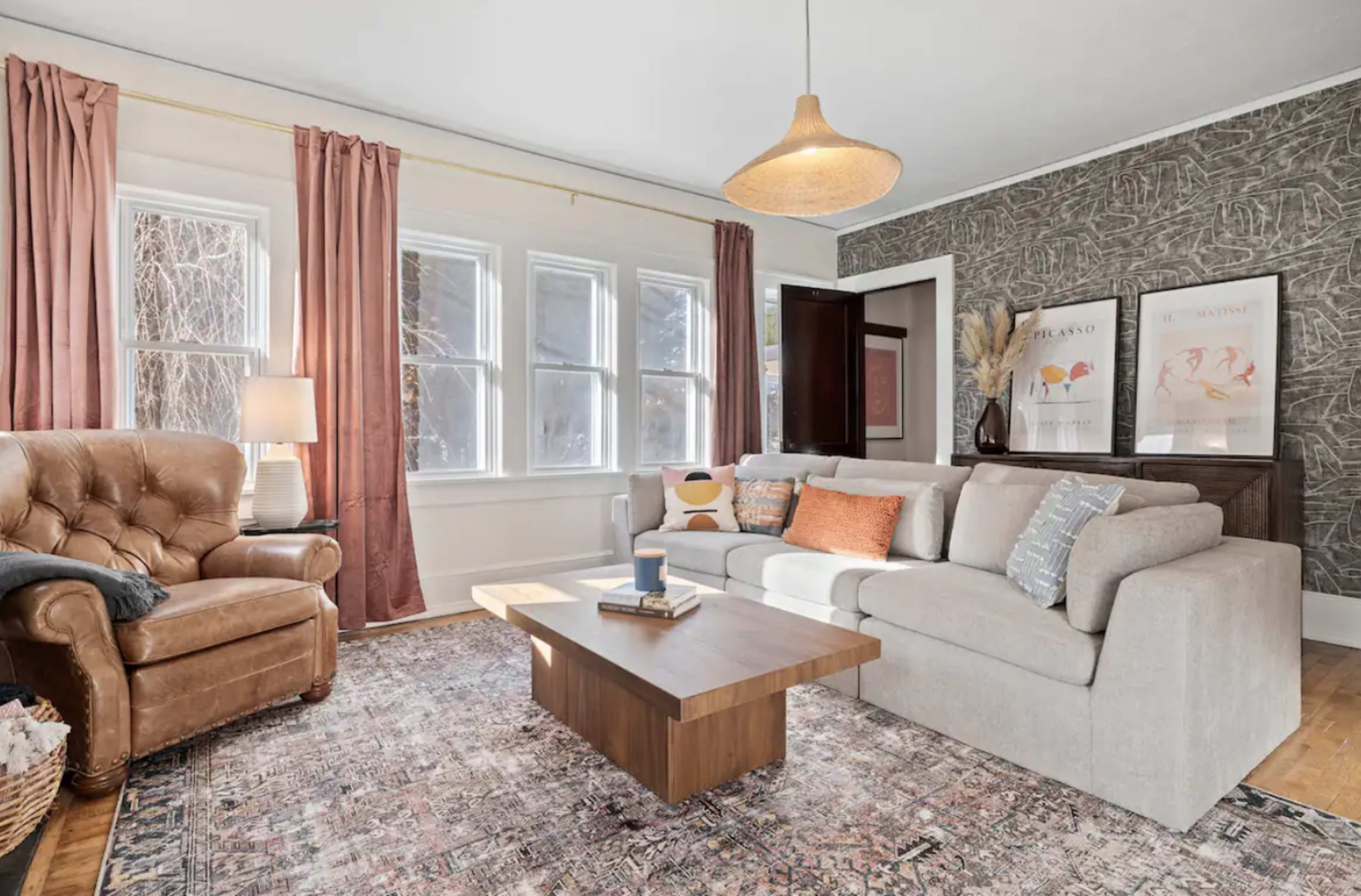 A cozy living room features a light gray sofa, a wooden coffee table, a brown armchair, and large windows adorned with light curtains.