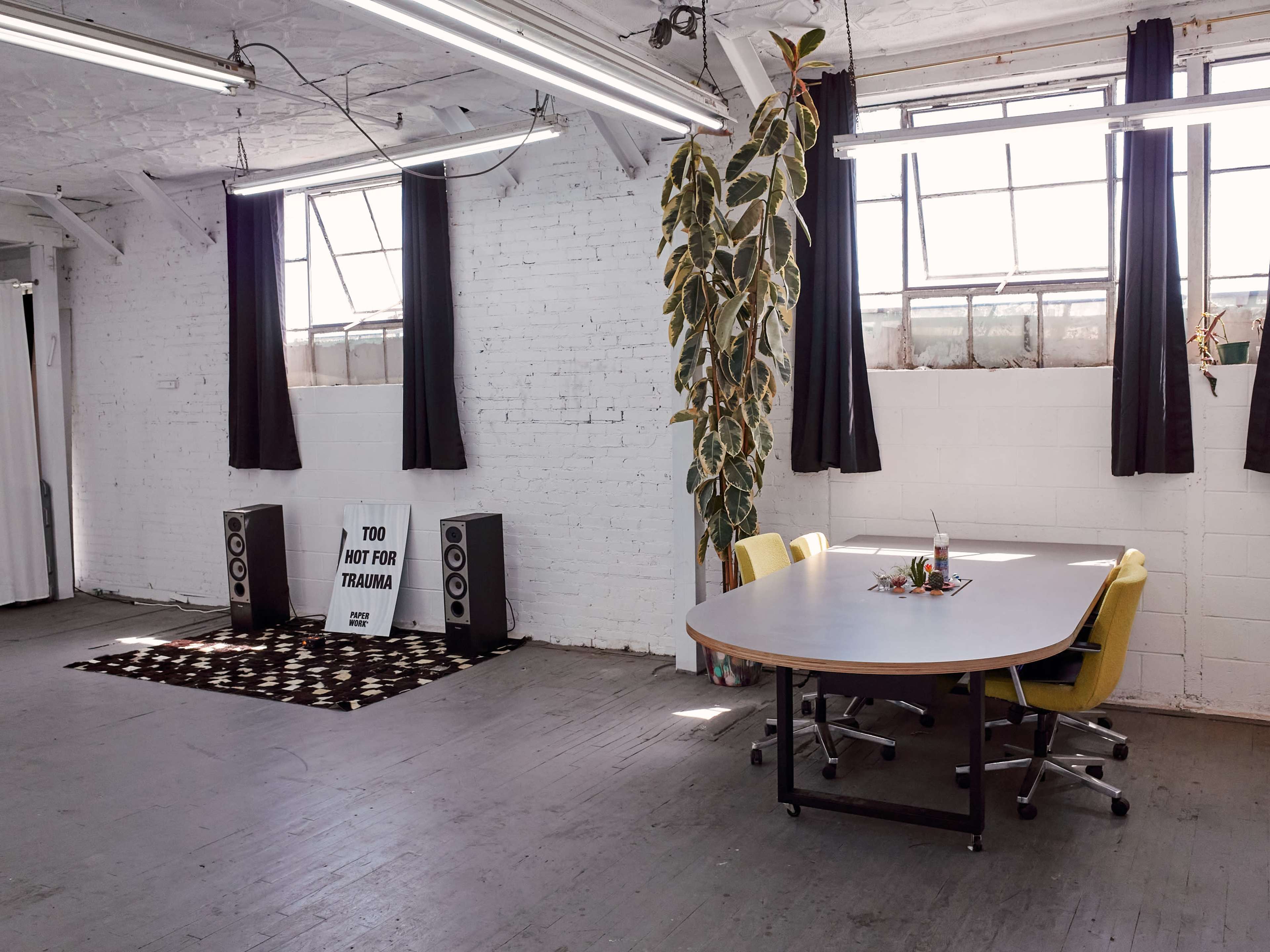 A spacious, well-lit room features a large circular table surrounded by yellow chairs, with a patterned rug, speakers, and a sign reading "TOO RAW FOR TRAUMA" in the background.