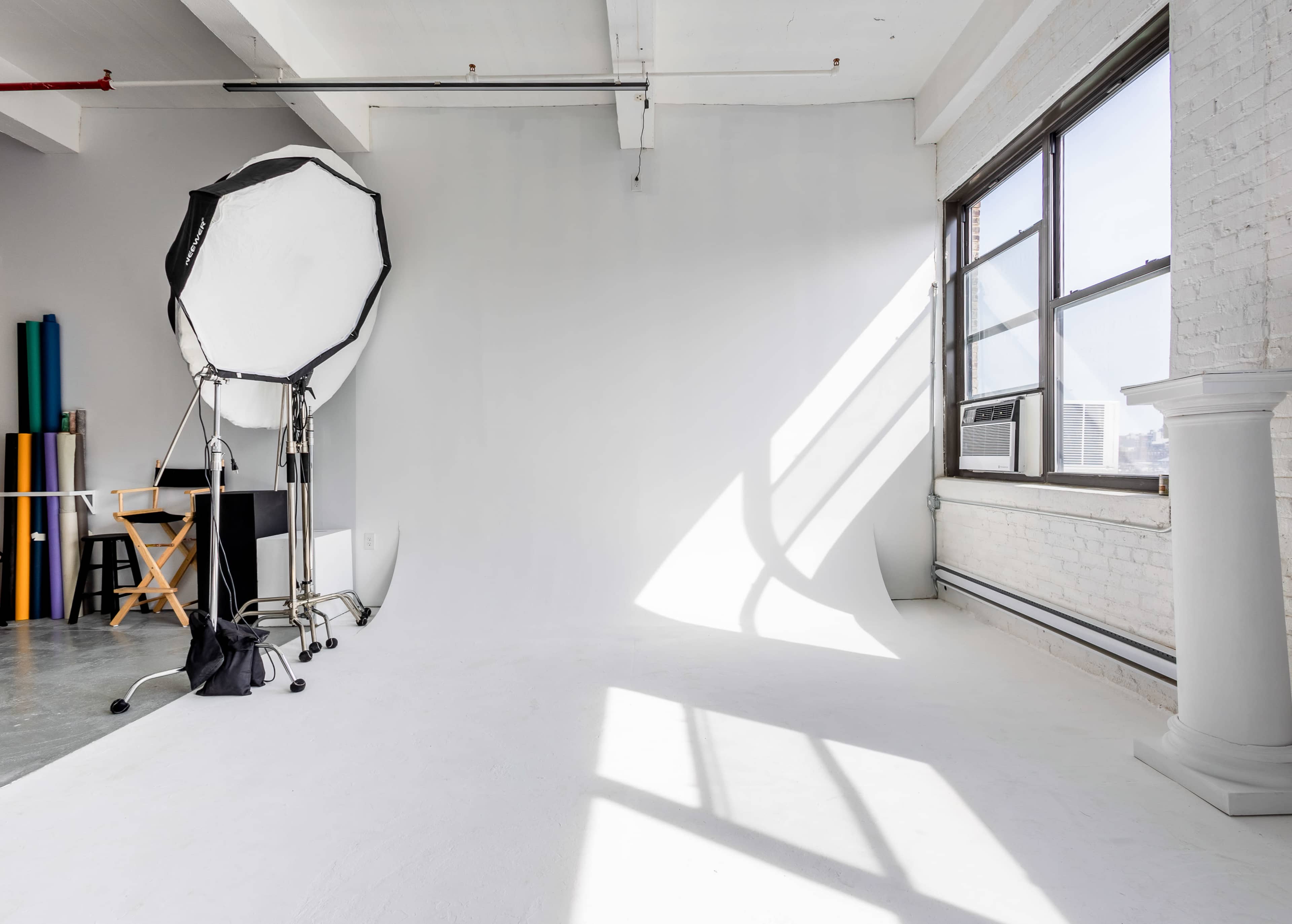 The image shows a photography studio with a white backdrop, a large umbrella softbox, and a window casting shadows on the floor.