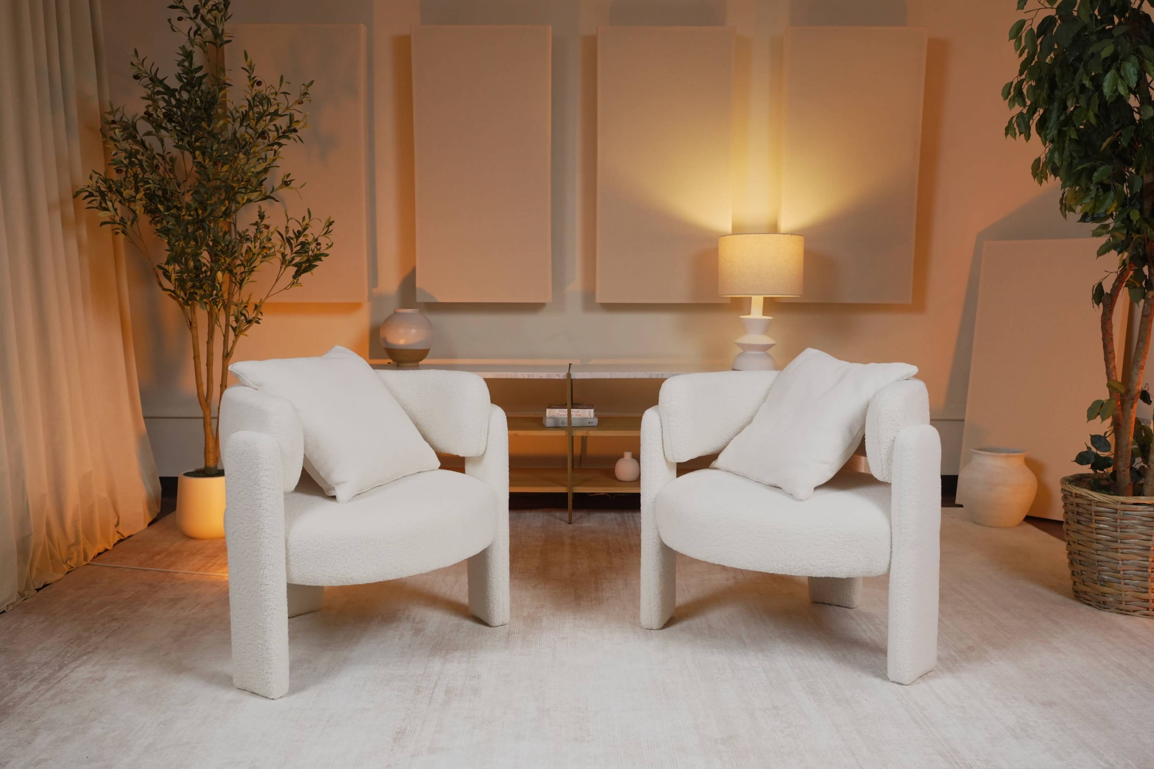 Two cream-colored chairs with rounded arms and soft cushions are positioned across from a glass coffee table in a softly lit room adorned with plants and wall panels.