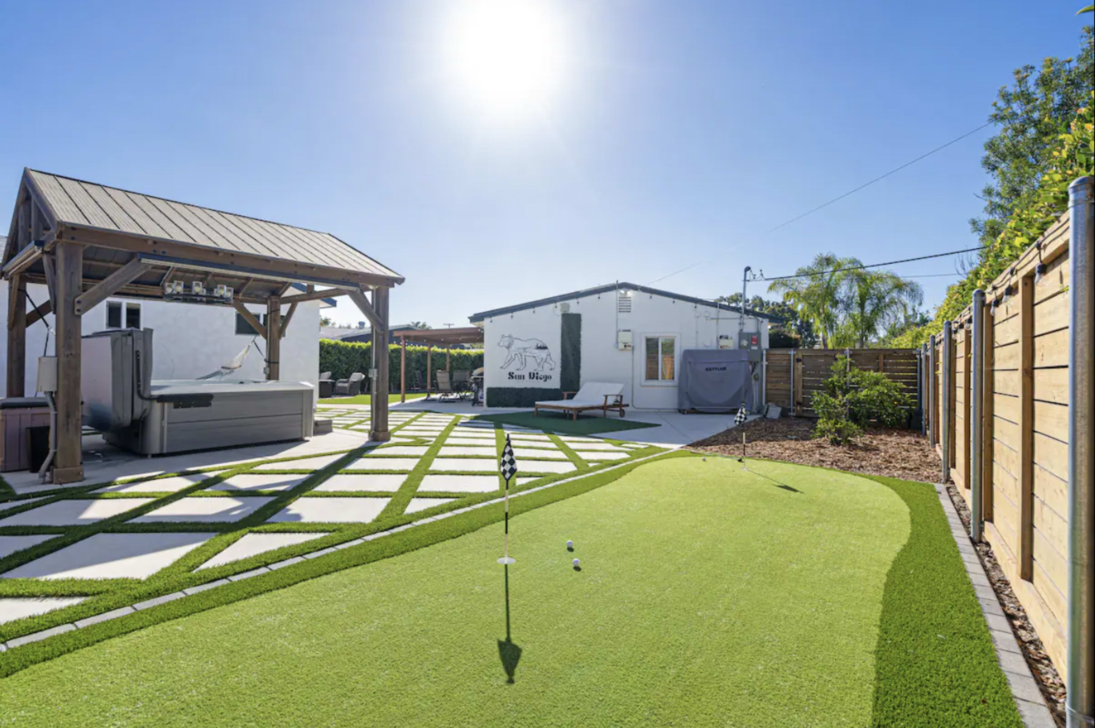 A backyard with a putting green, a spa under a wooden gazebo, and a modern white house surrounded by lush greenery.