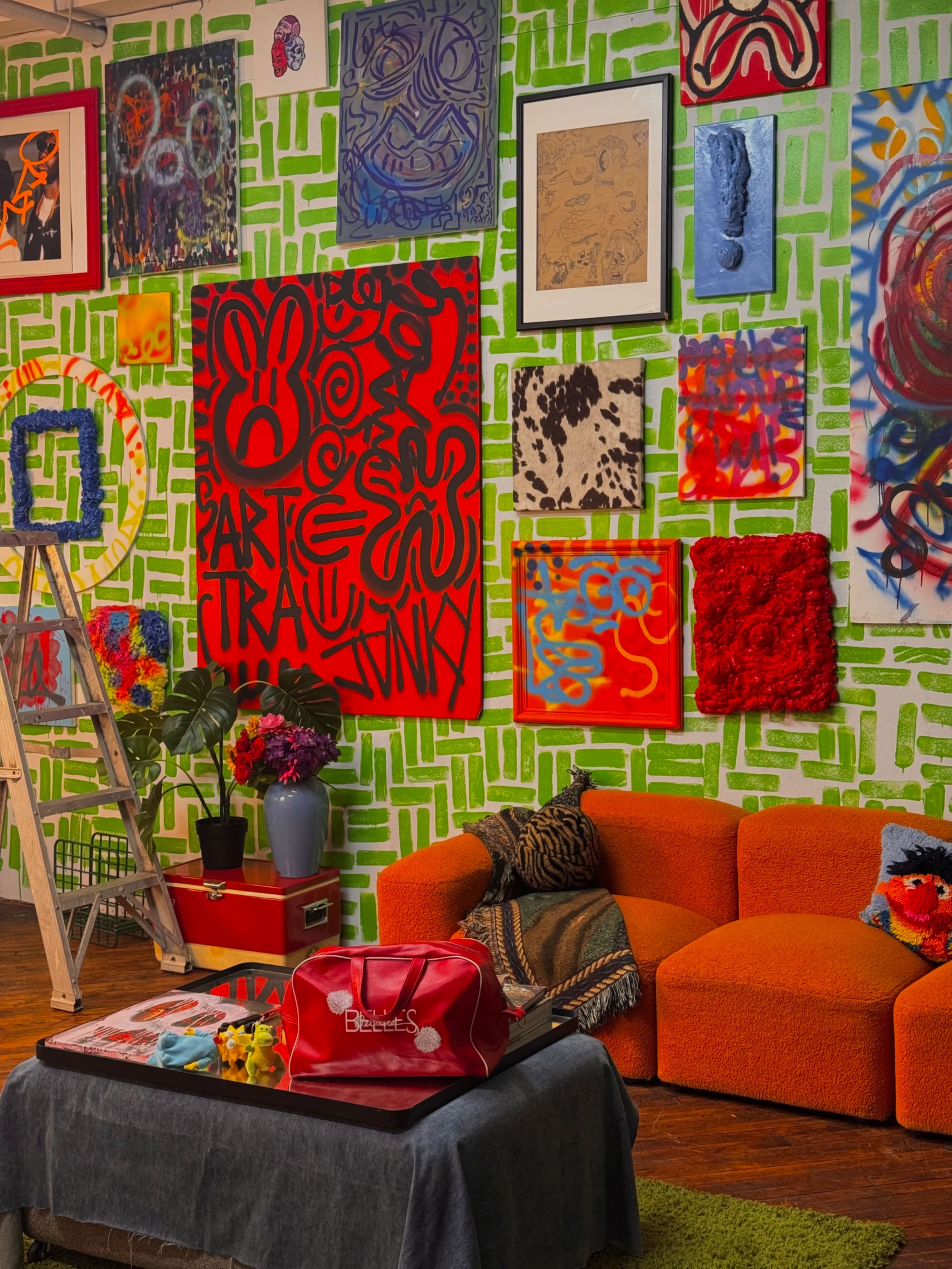 The image features a brightly decorated living space with an orange couch, a ladder leaning against a green and white patterned wall adorned with various colorful artworks.