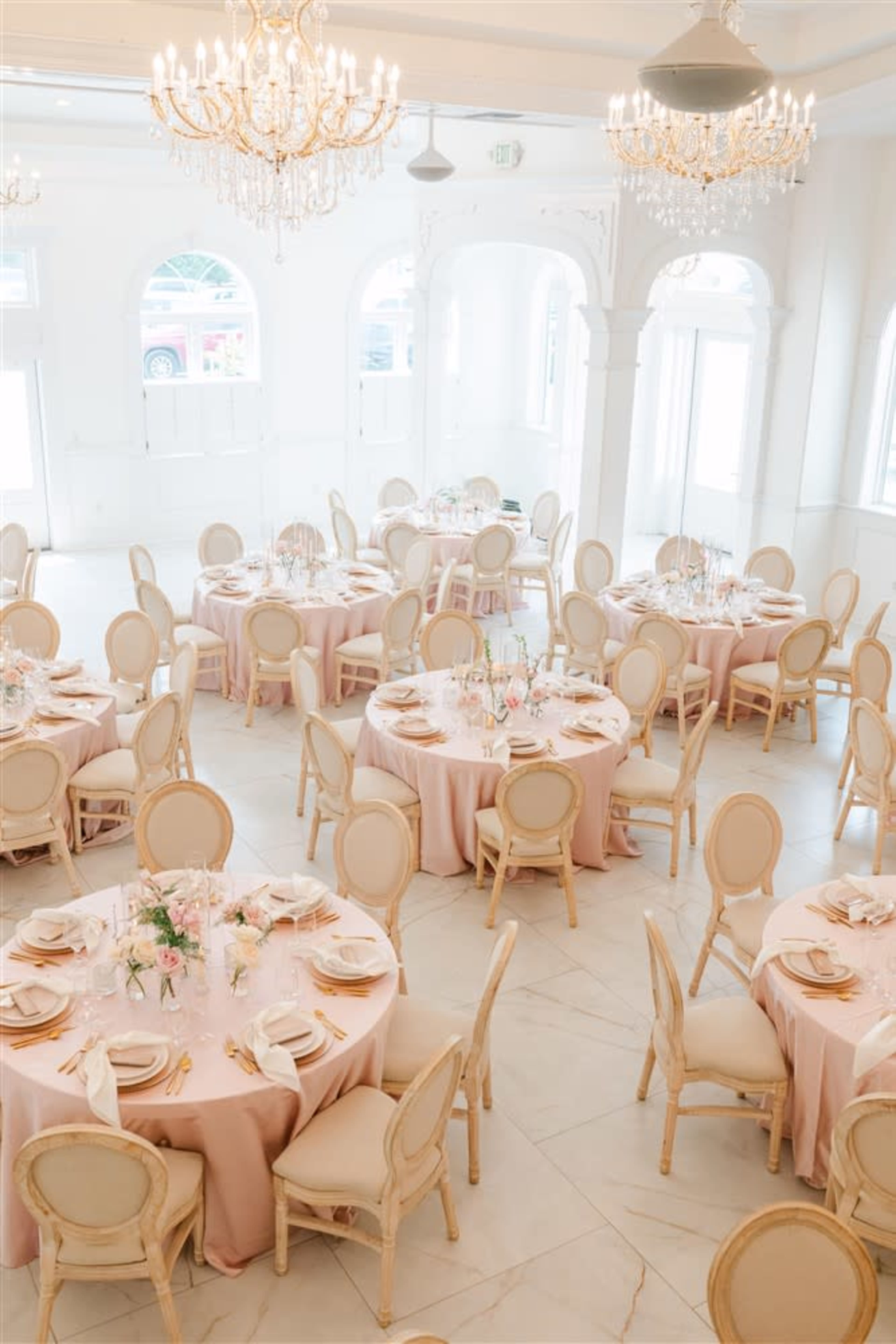 The image shows a banquet hall filled with multiple round tables, each set with pink tablecloths, dinnerware, and floral centerpieces, under elegant chandeliers.