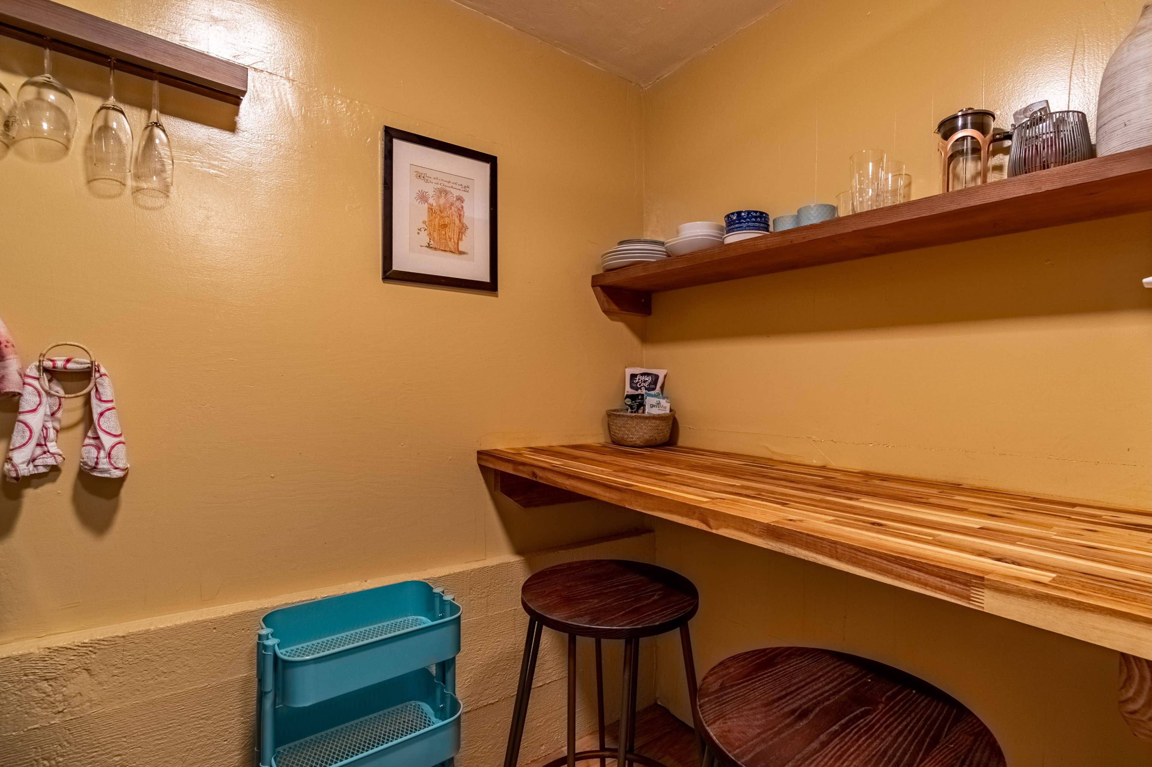 A small room with yellow walls, wooden shelves holding dishes and glassware, and a countertop made of wood, accompanied by two stools and a blue storage container.