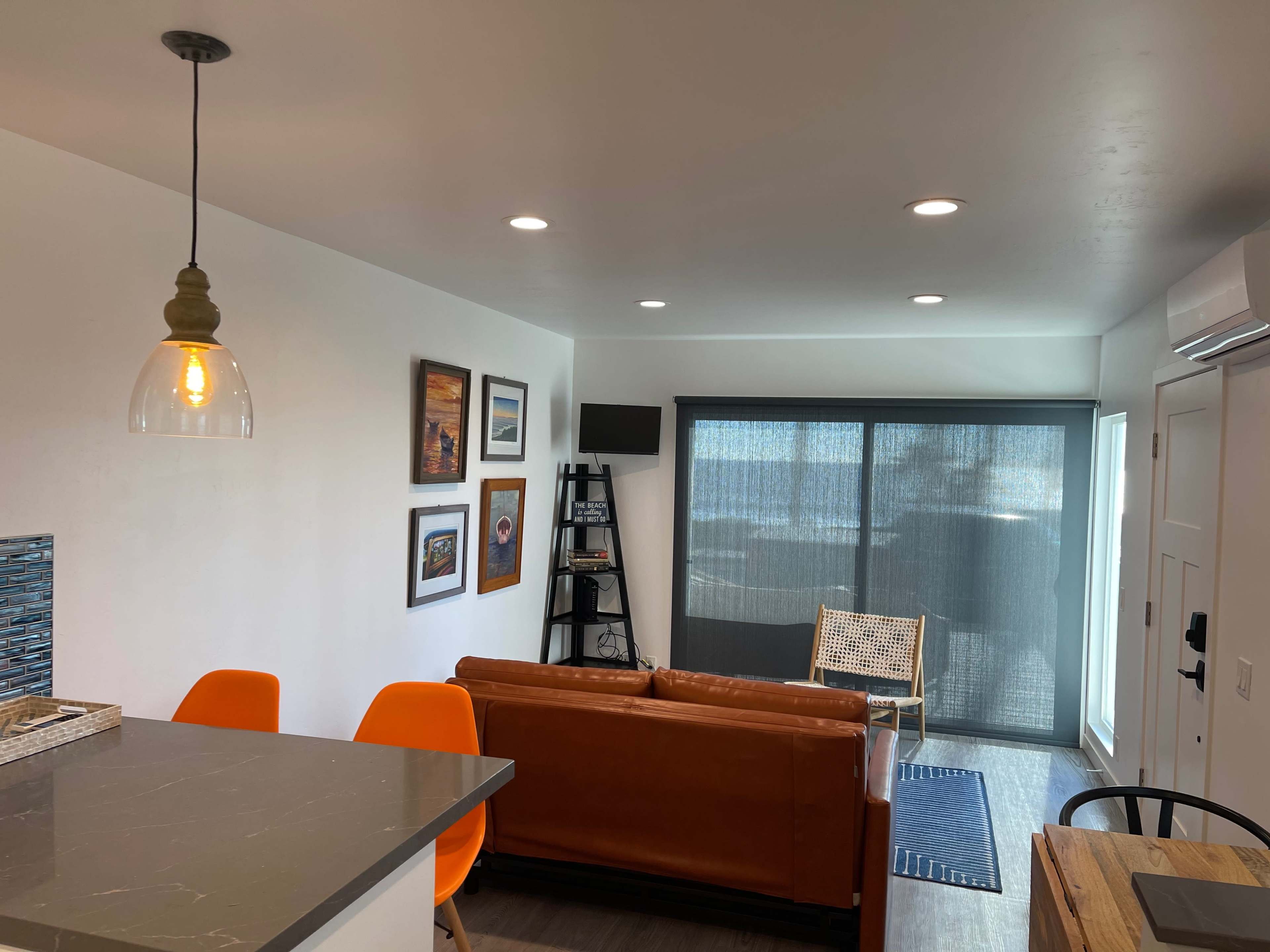 A modern living space features a brown leather sofa, a wooden dining table with orange chairs, and a sliding glass door covered by a black shade.
