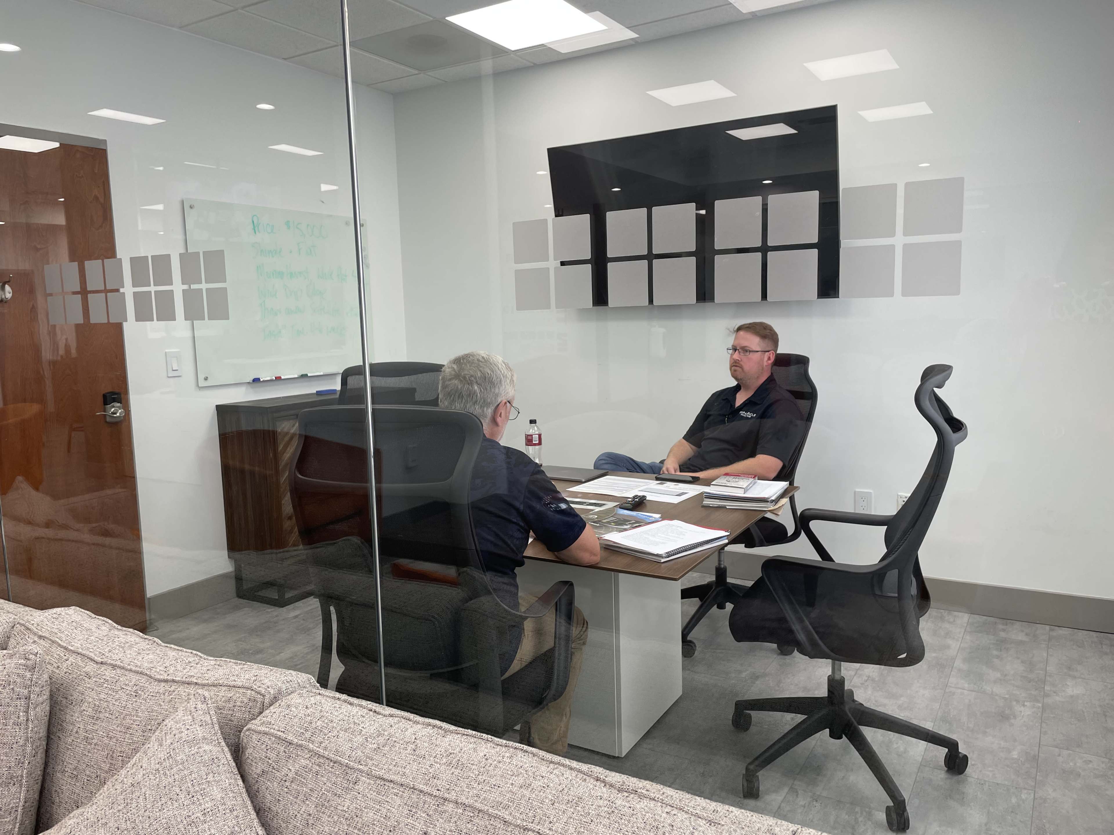 Two men are seated at a conference table in a glass-walled meeting room, discussing documents and notes.