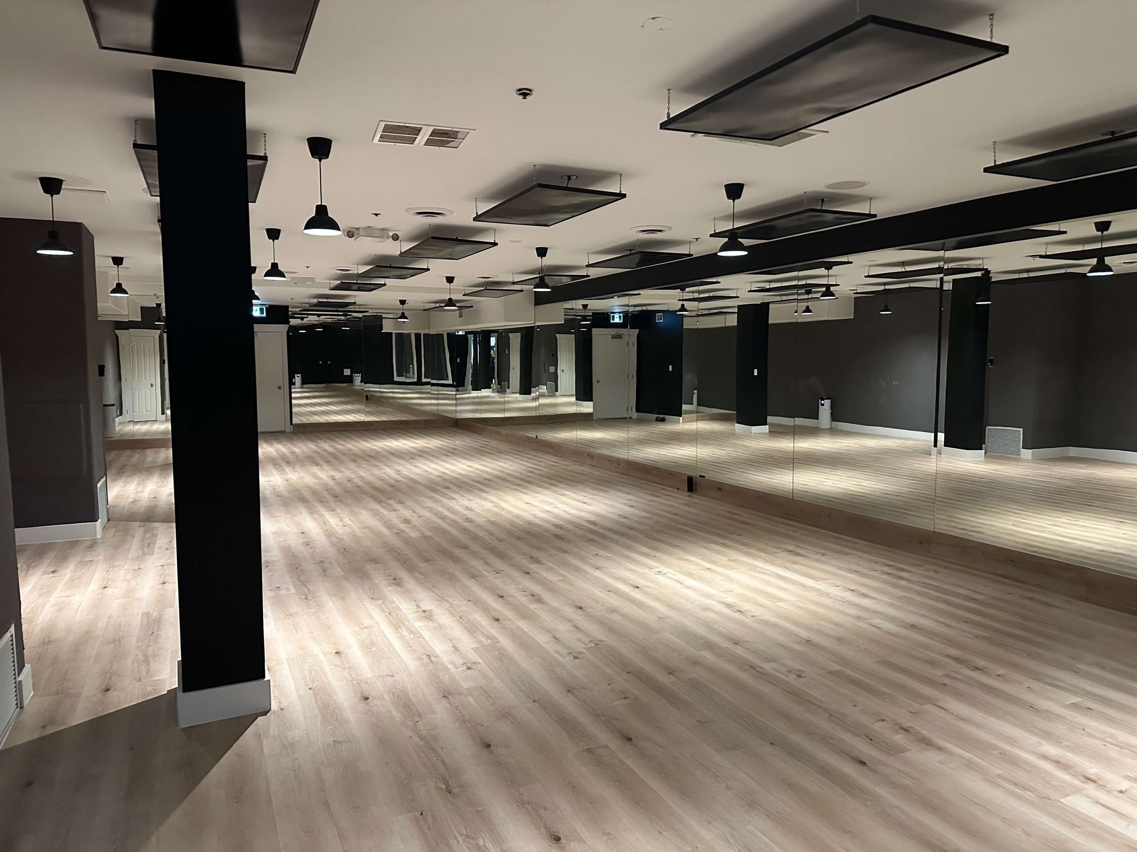 An empty mirrored studio with wooden flooring and overhead lights.