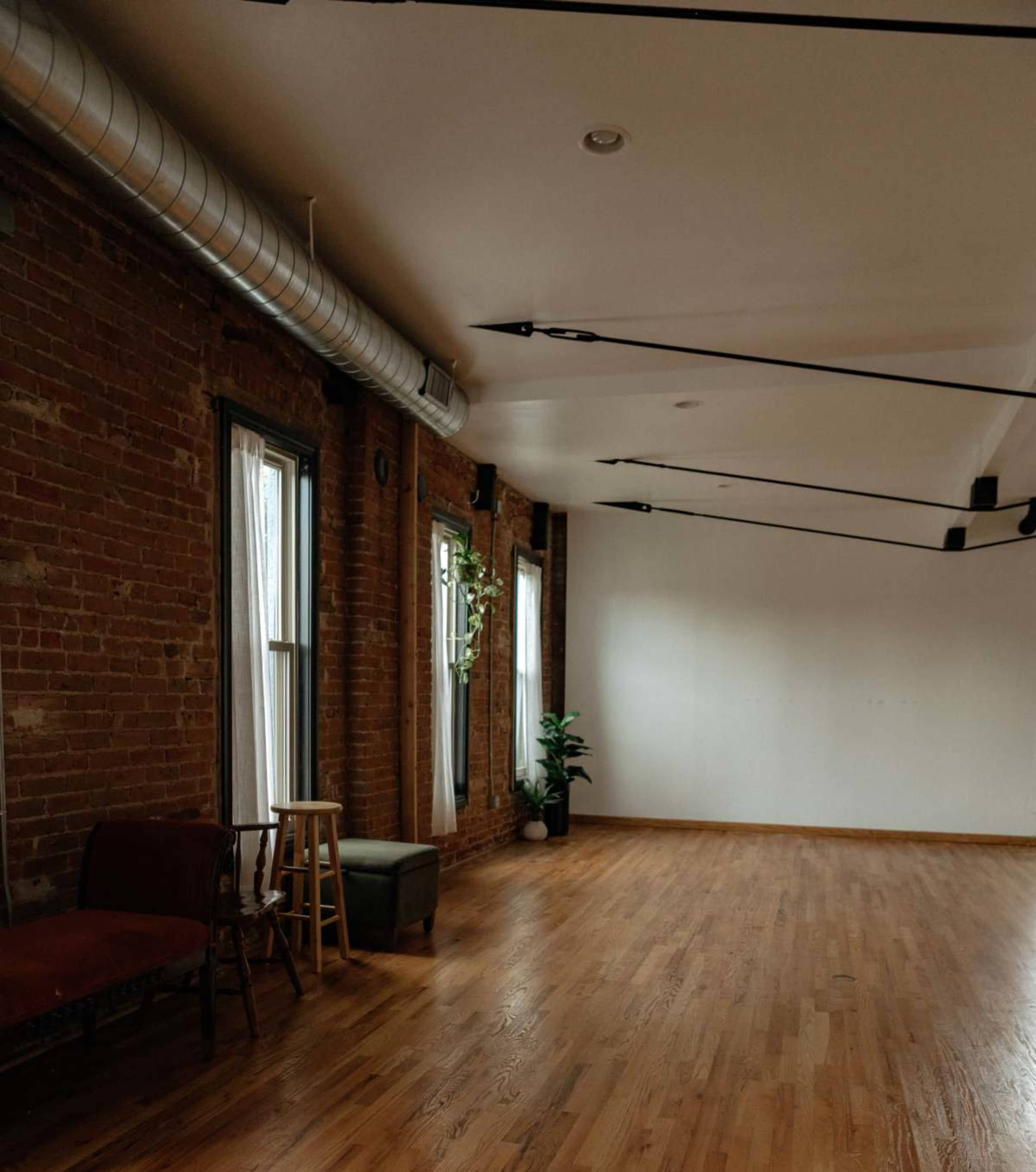 The image shows an empty, well-lit room with exposed brick walls, large windows, and wooden flooring, featuring minimal furniture including a stool and a small plant.