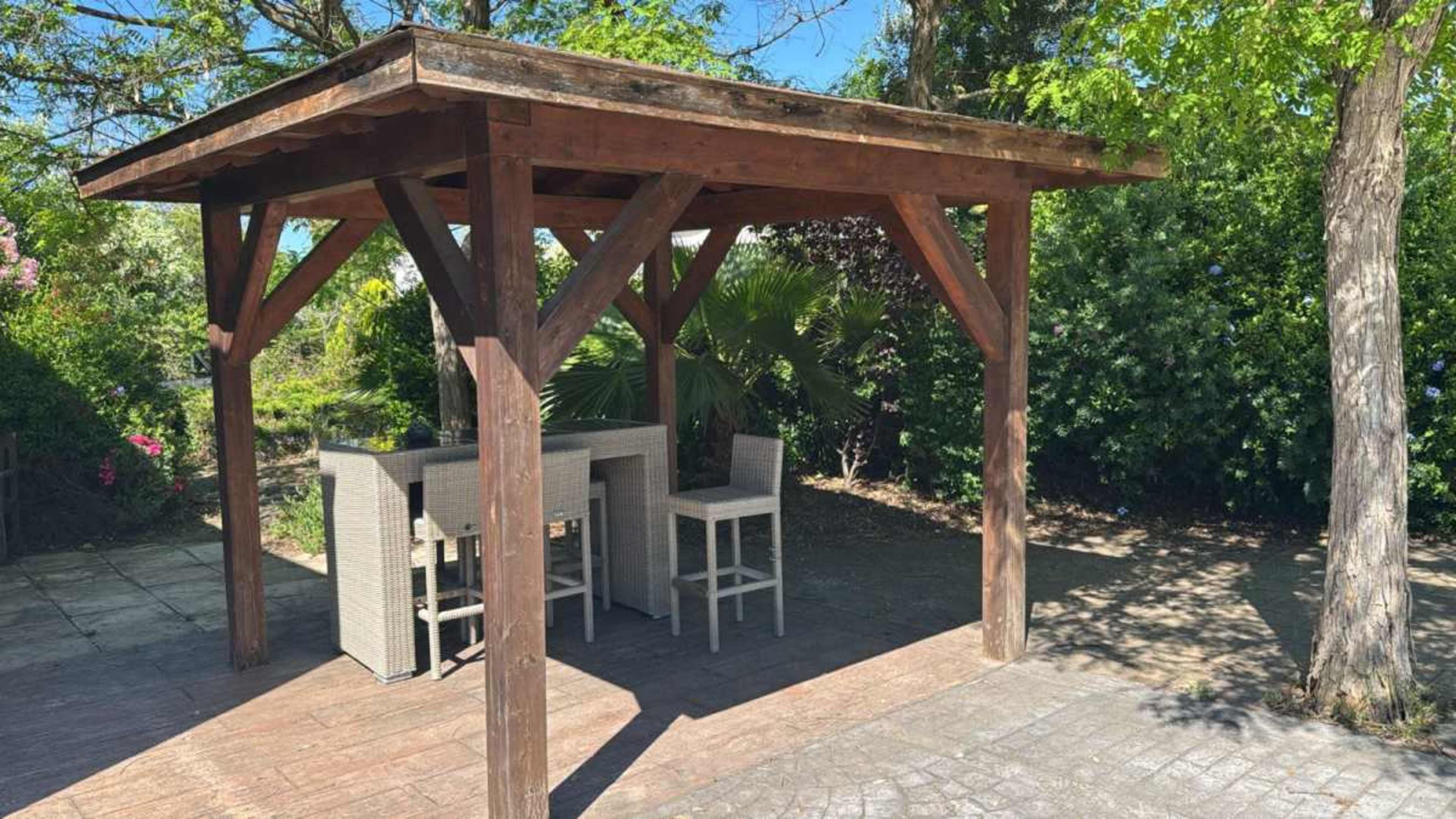 A wooden gazebo with a bar-style seating area is situated in a landscaped outdoor space.