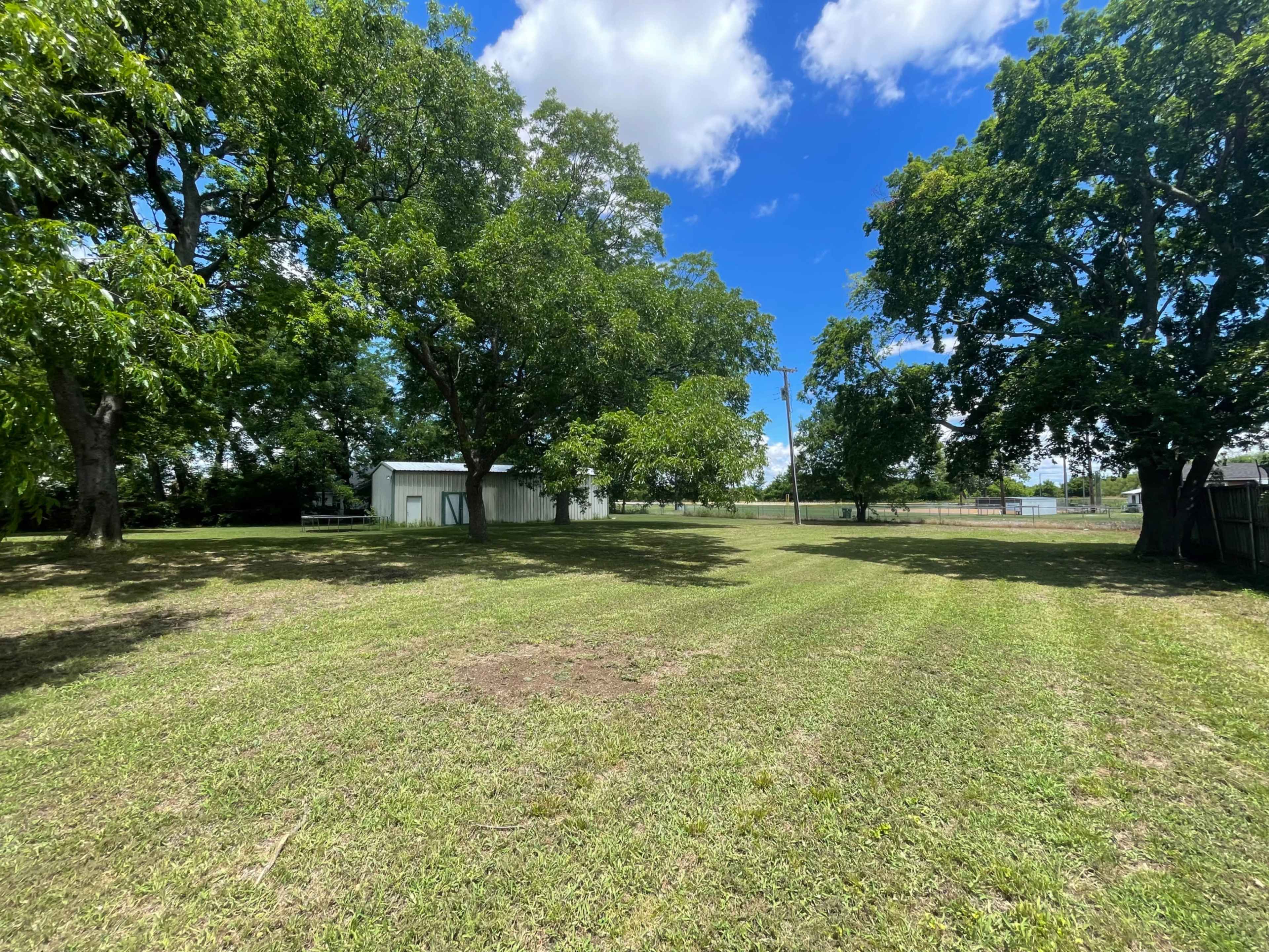 The image shows an open grassy area with several large trees and a small white building in the background under a partly cloudy blue sky.