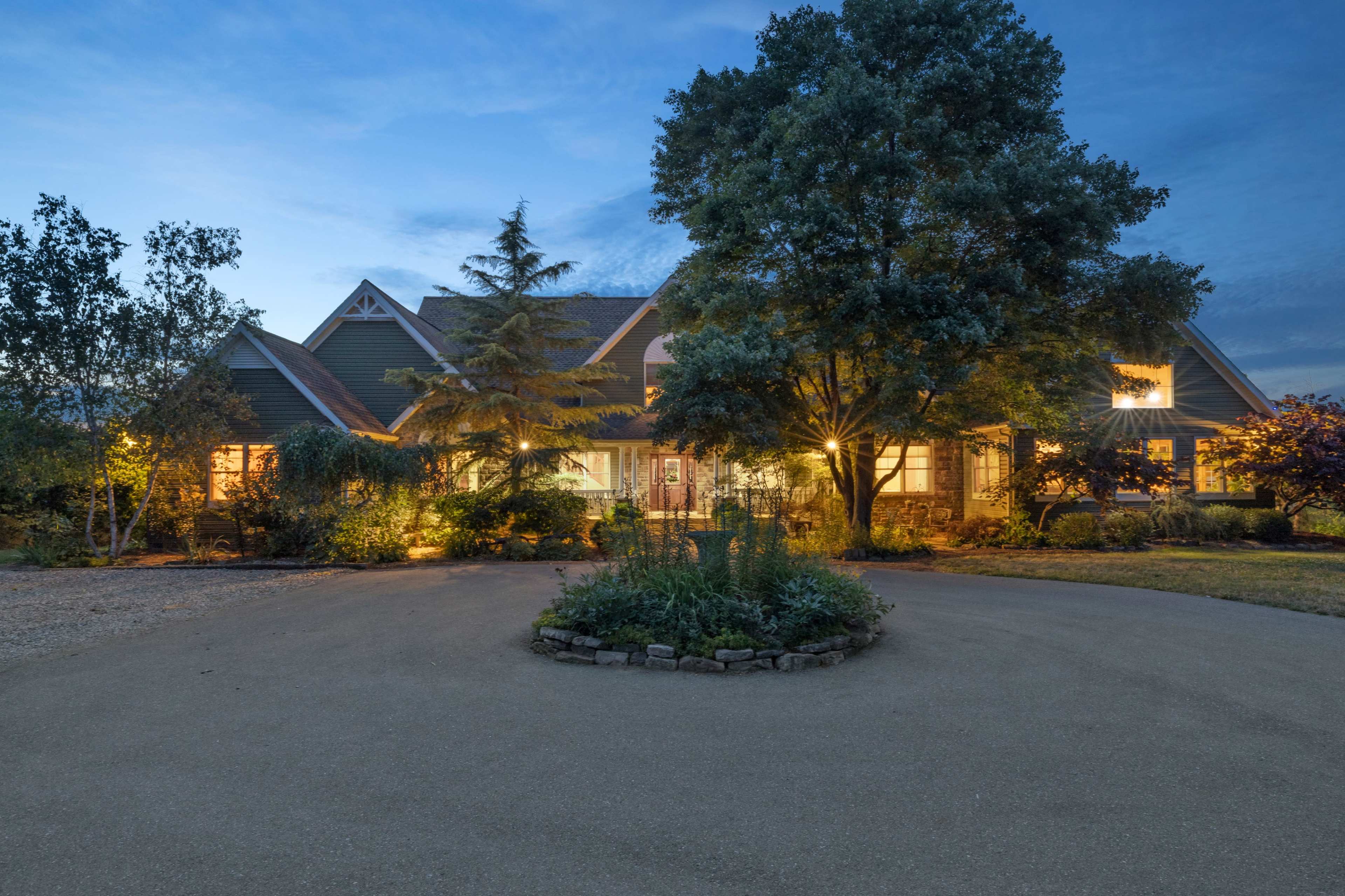 A spacious house with a landscaped front yard is illuminated at dusk, featuring a circular driveway and various trees.