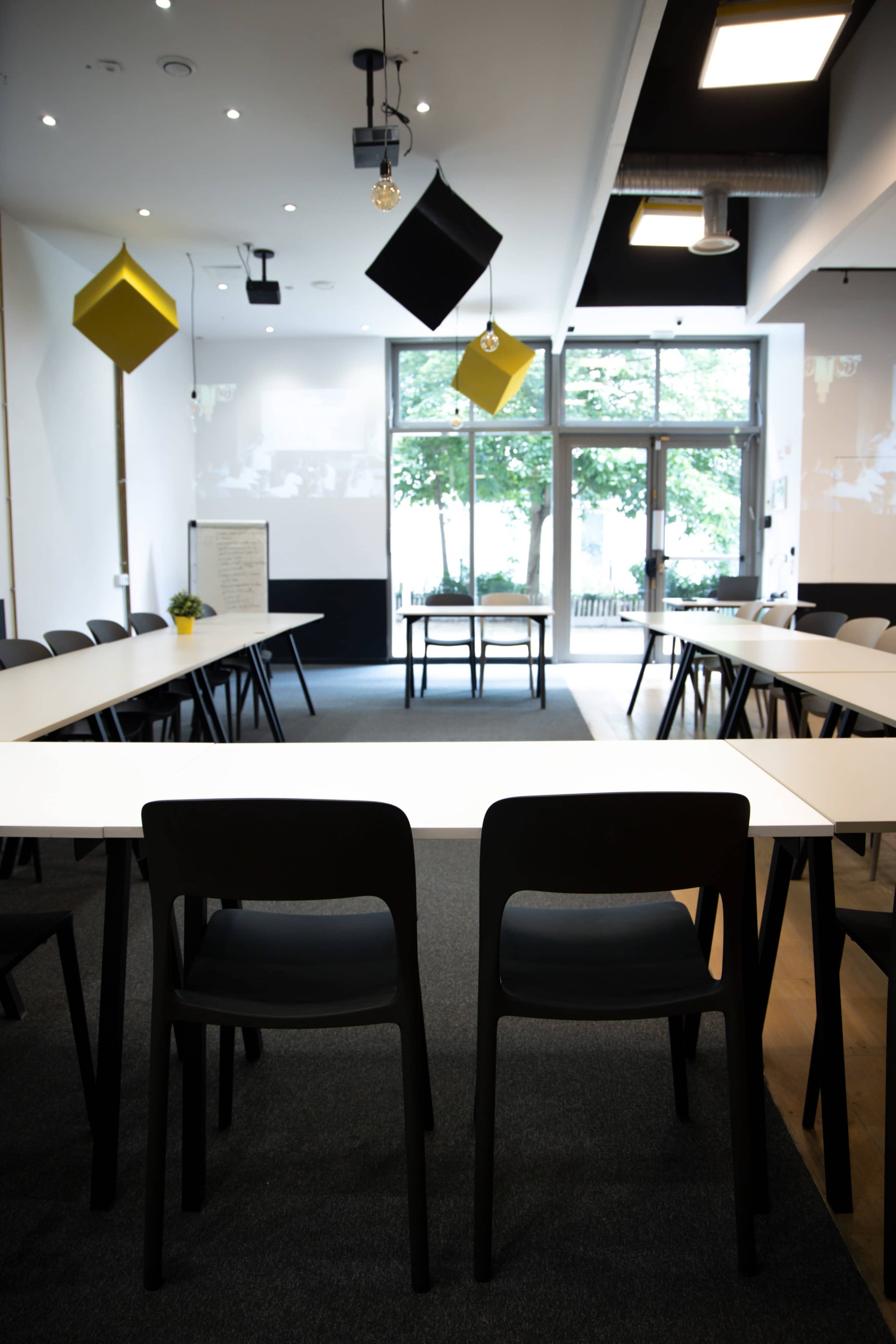 A modern meeting room features several white tables arranged in a U-shape, with black chairs and geometric decorations hanging from the ceiling.
