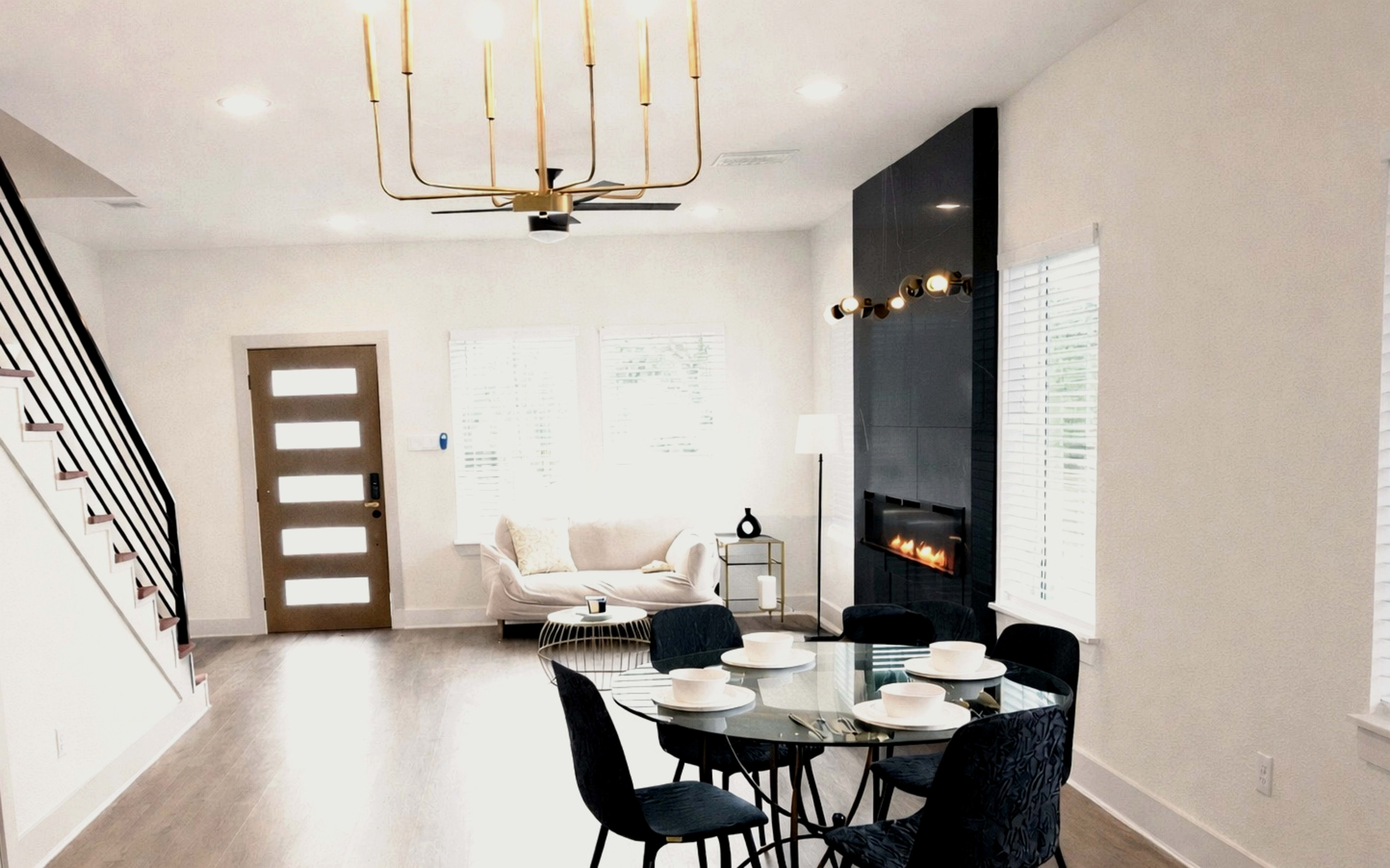 The image shows a modern living space with a dining area featuring a glass table and black chairs, adjacent to a seating area with a sofa and a wall-mounted electric fireplace.